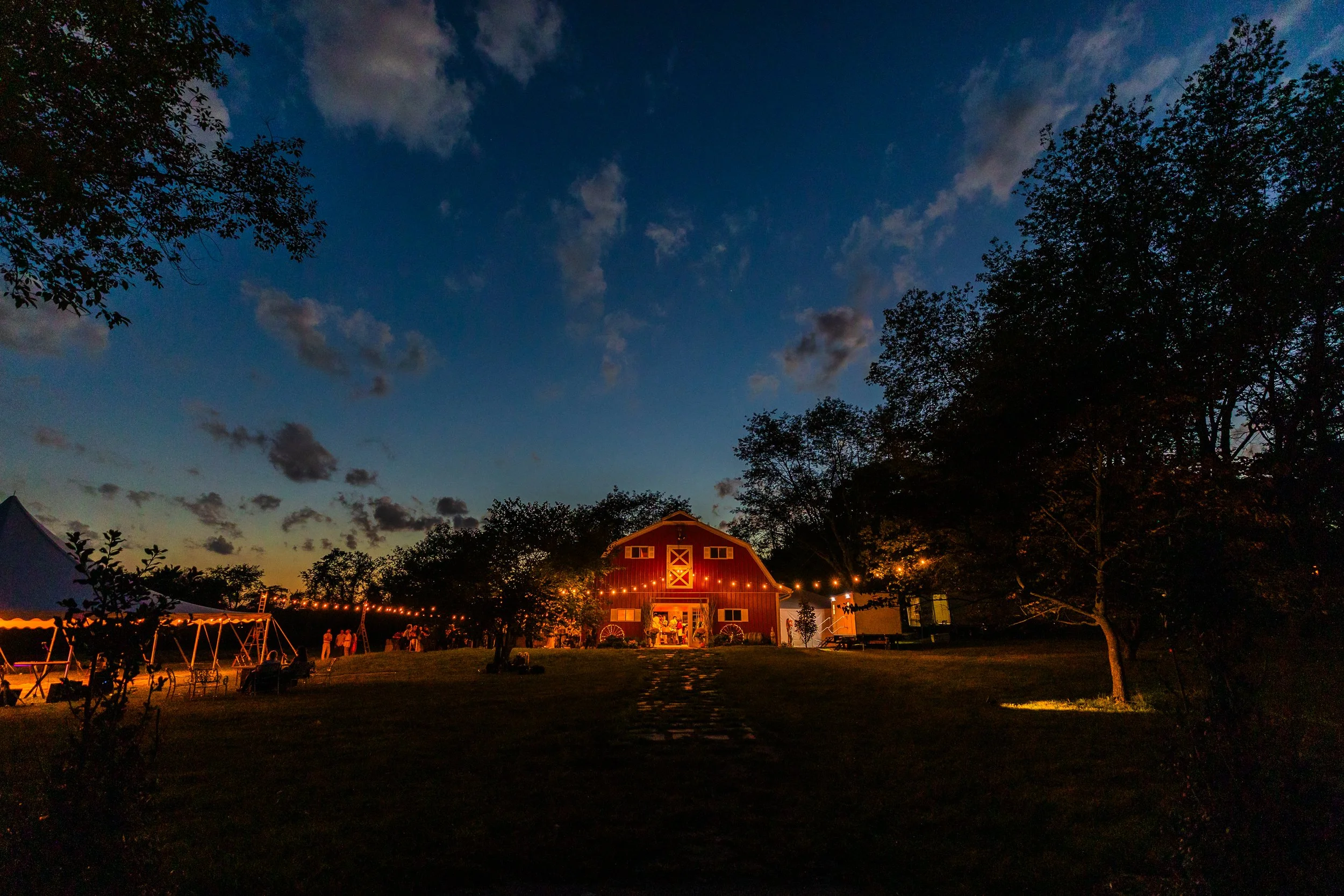 A large red barn decorated with string lights in a rural setting during twilight, with trees on either side and a cloudy sky overhead.