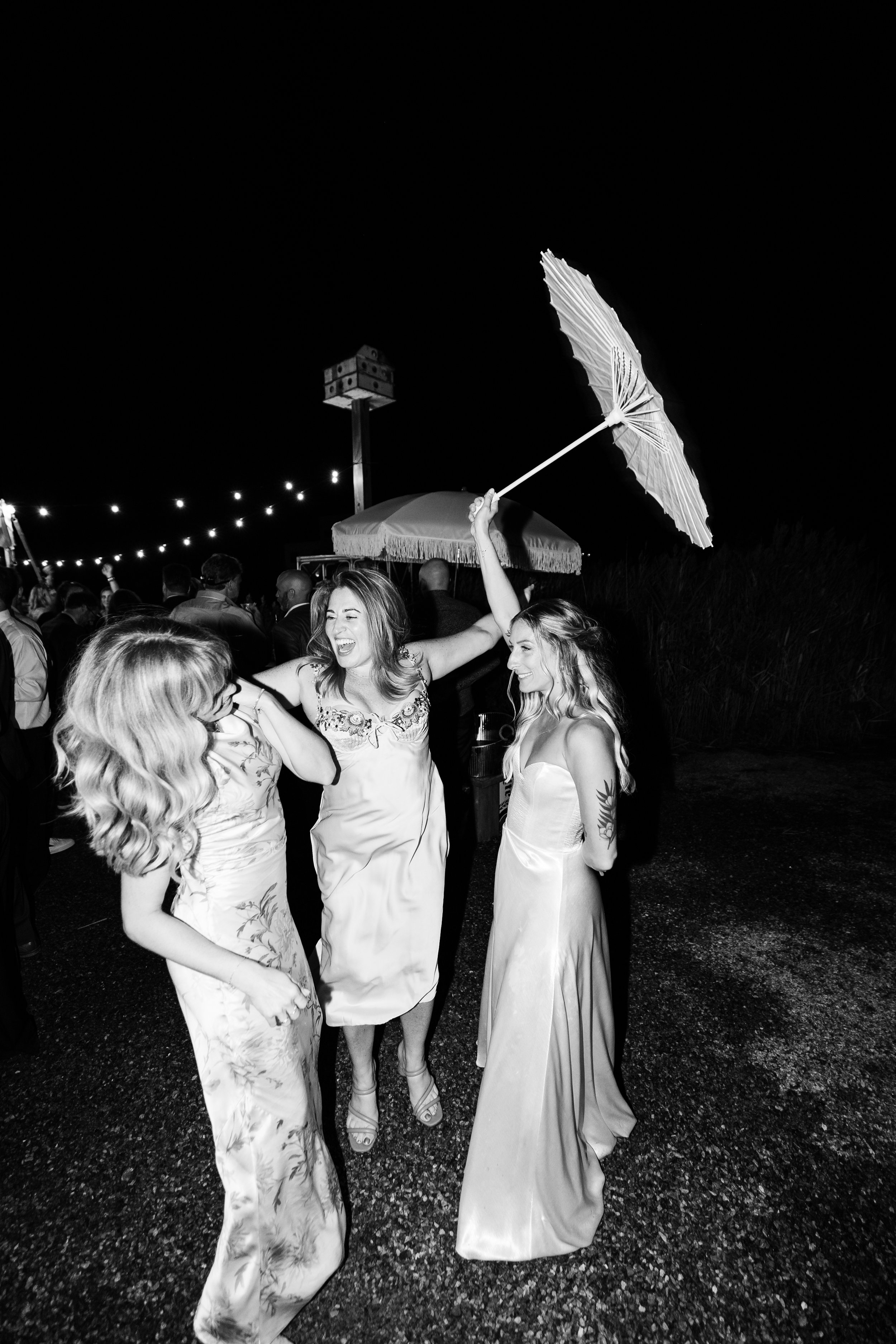 Three women celebrating at a wedding reception outdoors at night, with one holding a parasol above their heads.