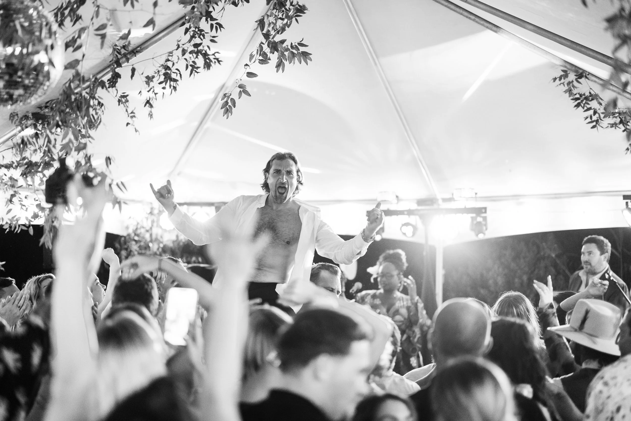 A man with long hair and a beard, wearing an open shirt and no shirt underneath, stands on a dance floor with arms raised and mouth open, appearing to sing or shout at a lively party under a canopy tent, surrounded by a crowd of people taking photos 