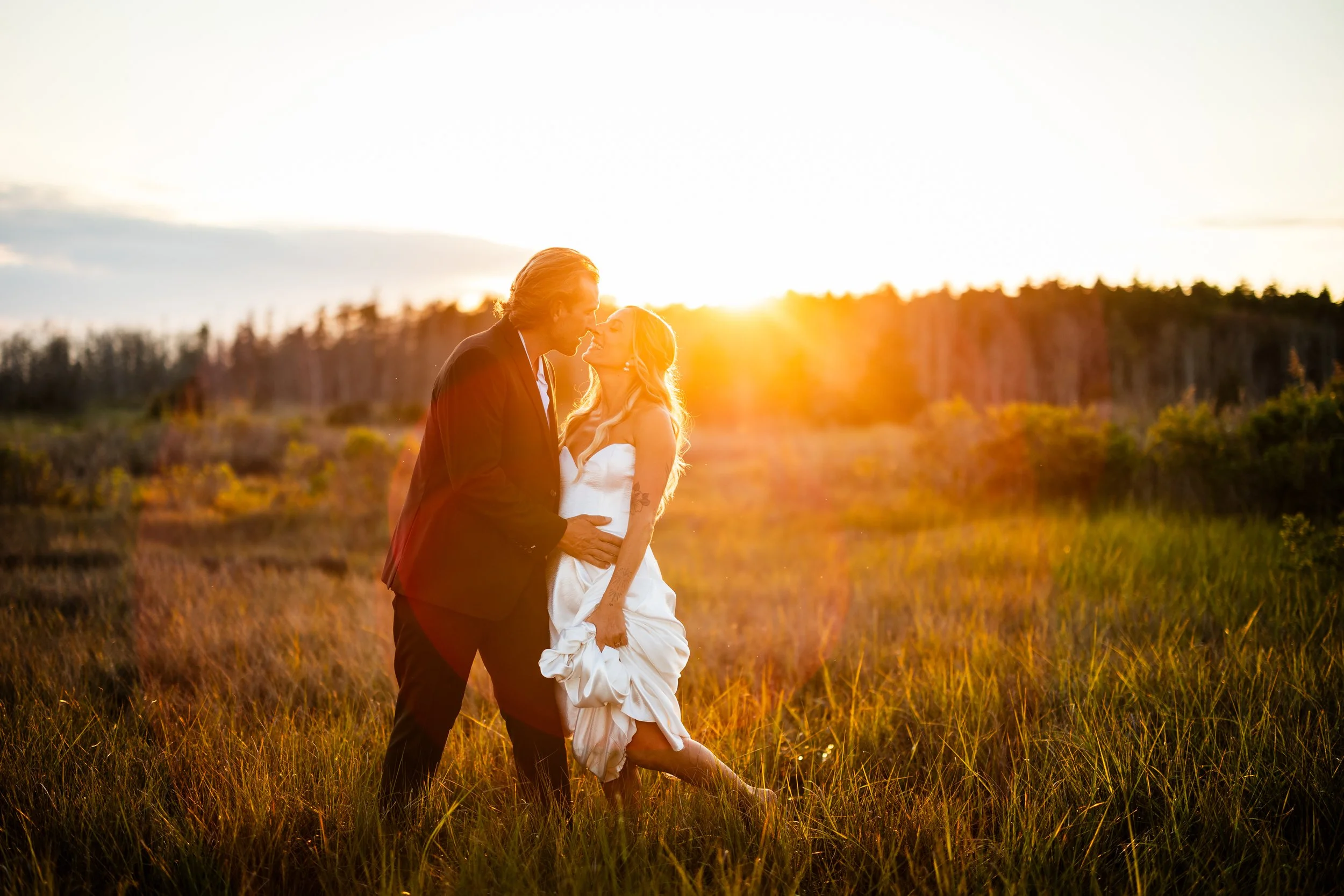 A couple dressed in wedding attire standing in a grassy field during sunset, gazing at each other lovingly.