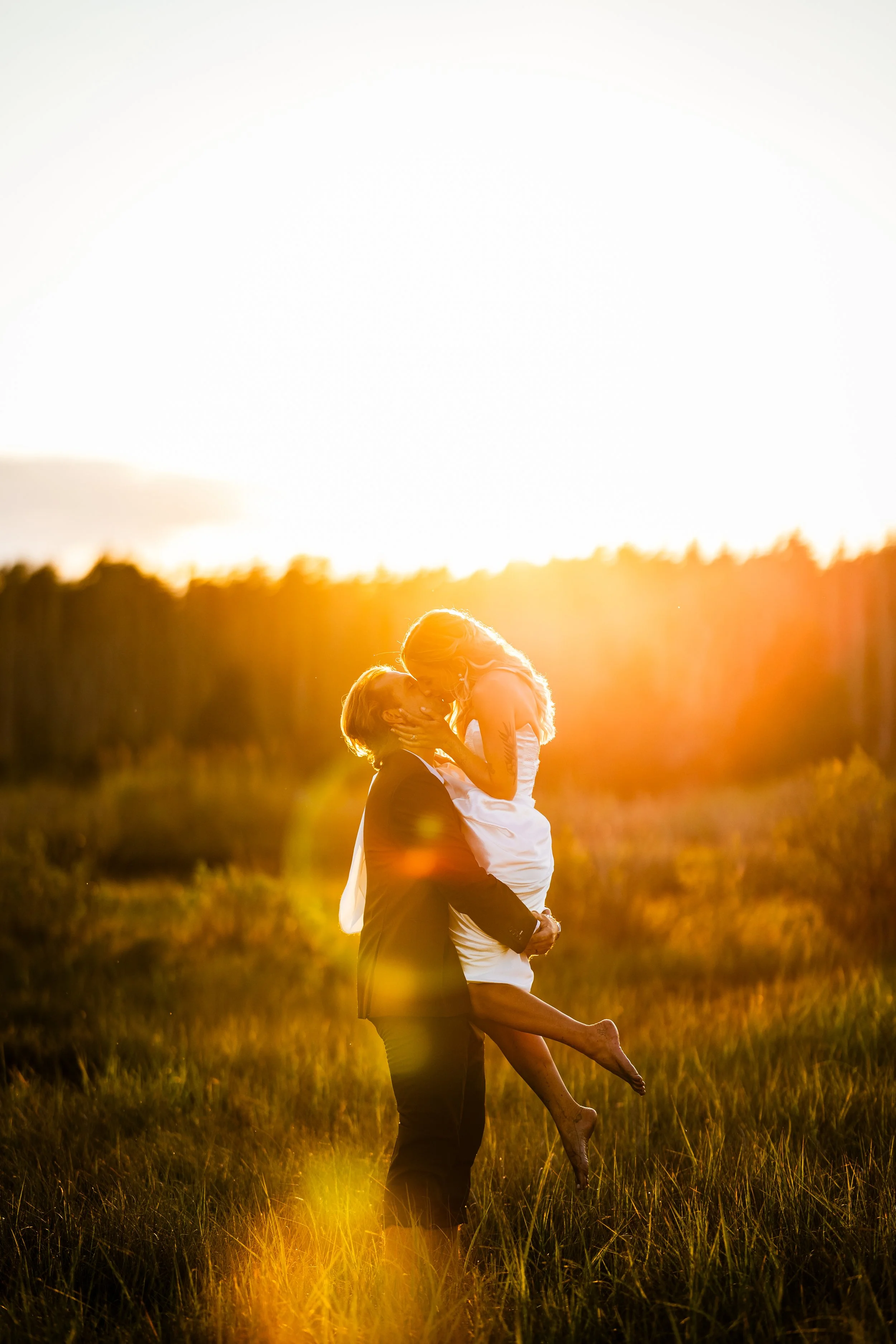 A man in a suit and a woman in a white dress embrace in a field at sunset, with the man lifting the woman off the ground as they share a kiss.