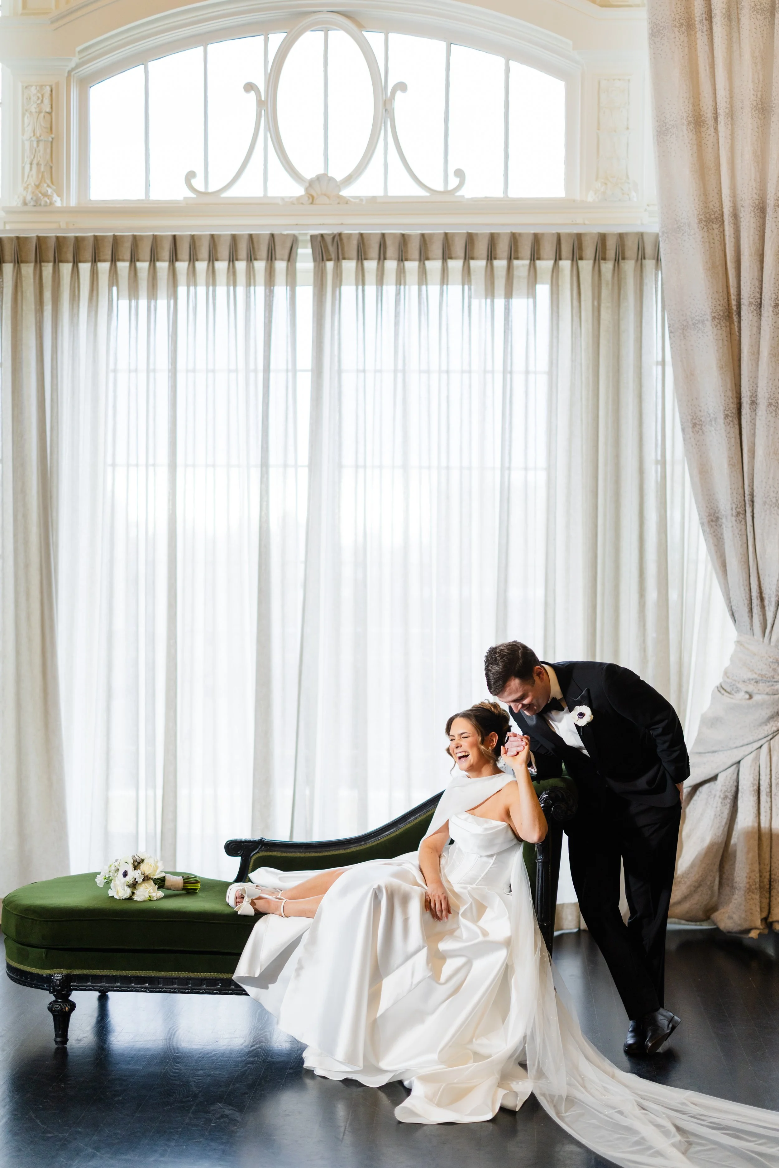 A bride in a white wedding dress and a groom in a black tuxedo are laughing together in a bright room with large windows and white curtains. The bride is sitting on a vintage green sofa, and the groom is leaning over her, holding her hand.