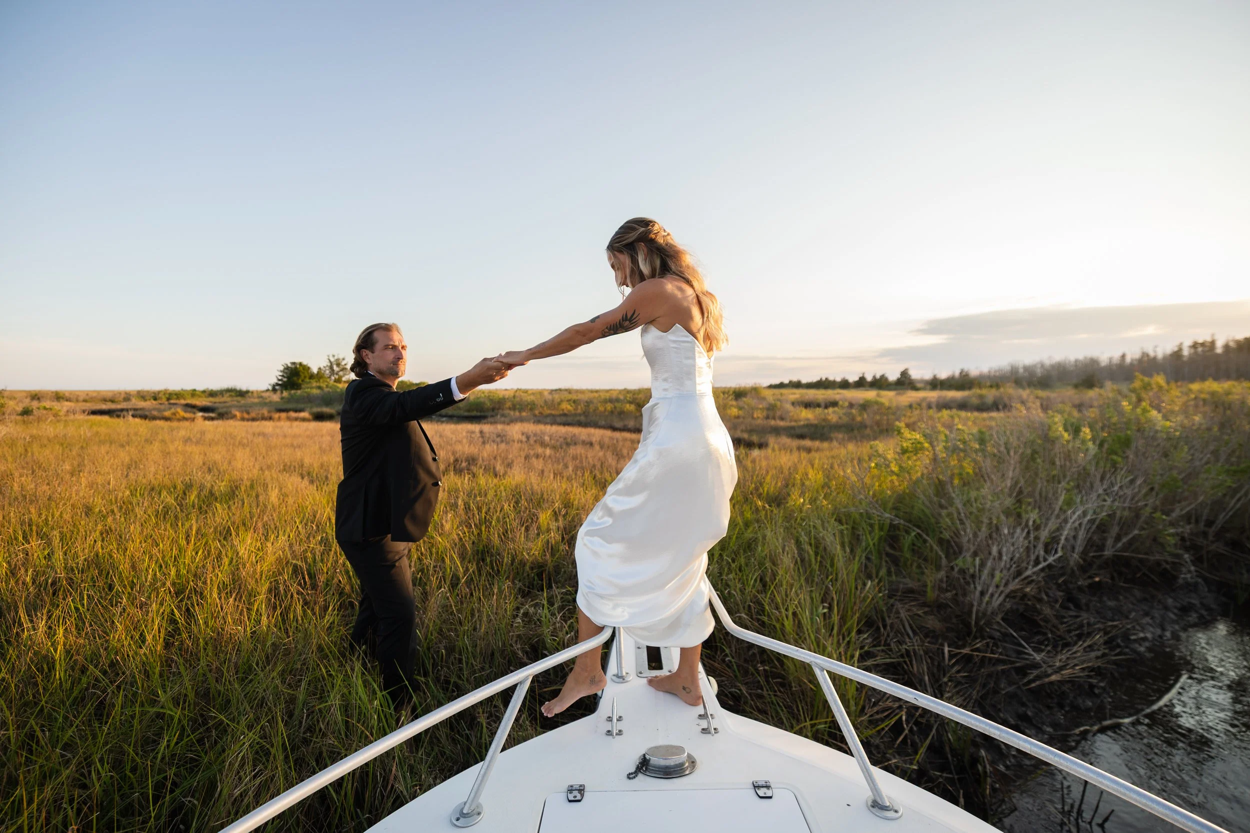 A bride standing on the bow of a boat, reaching out to hold hands with a groom standing on a grassy marshland at sunset.