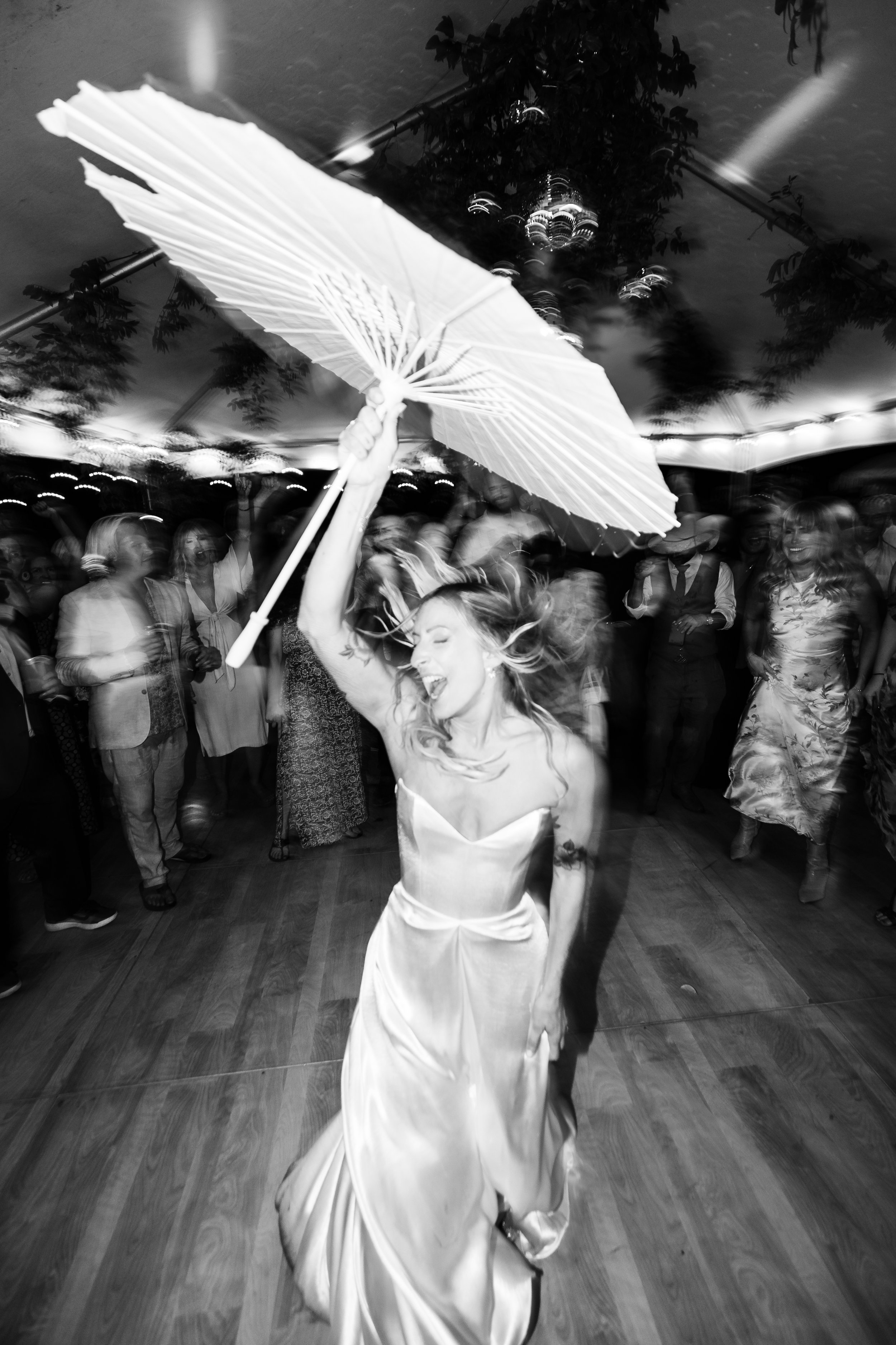 A woman in a wedding dress holding a parasol and dancing at a celebration or party, with several people in the background.