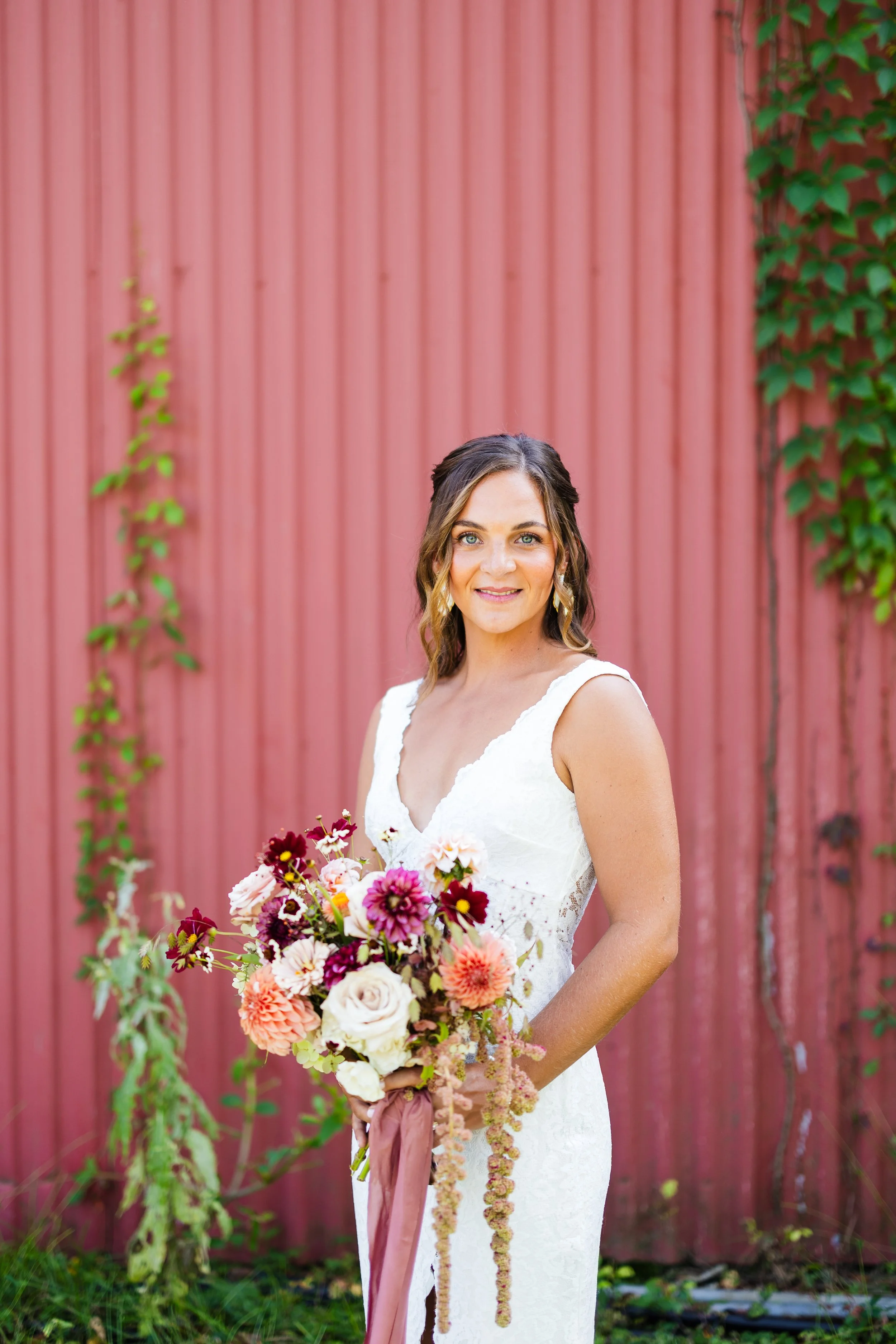 Bride in a white dress holding a bouquet of flowers, standing outdoors in front of a pink wooden wall with green climbing vines.