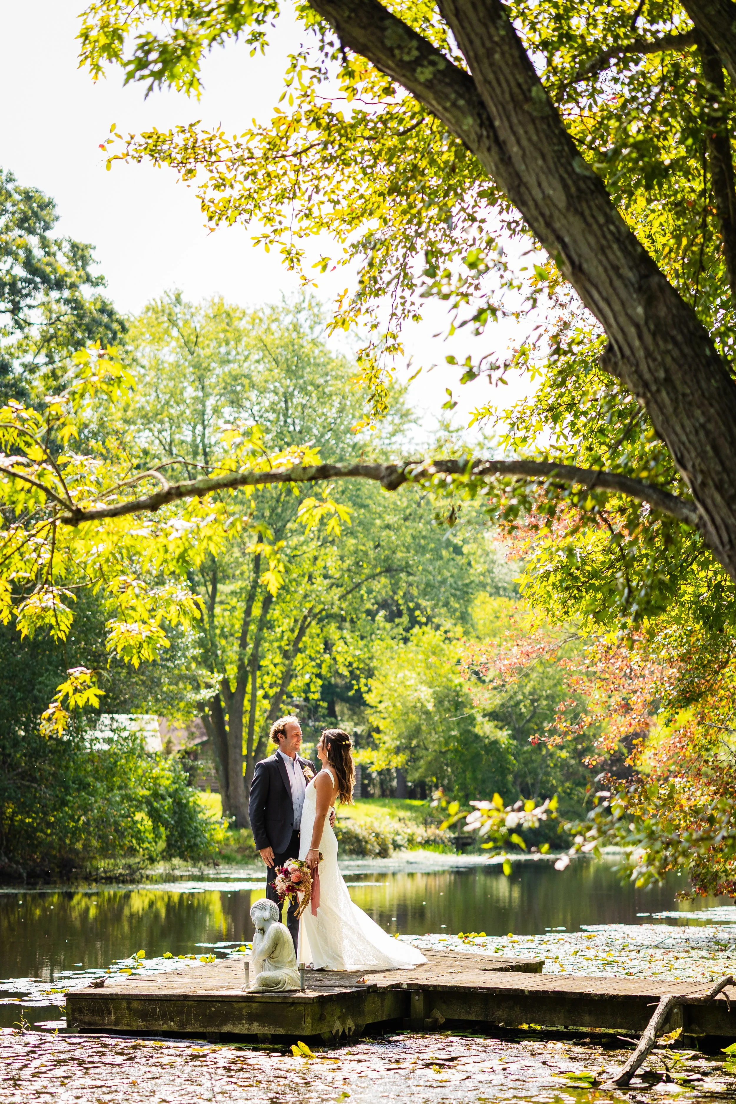 A bride and groom standing on a wooden dock by a pond, surrounded by lush green trees, with the bride holding a bouquet, during daytime.