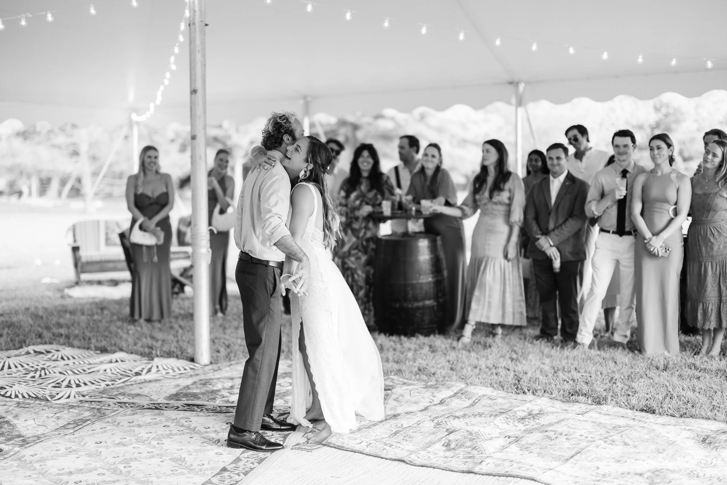 Black and white photo of a wedding reception, with a newlywed couple sharing a kiss or dance under a tent, surrounded by friends and family watching.