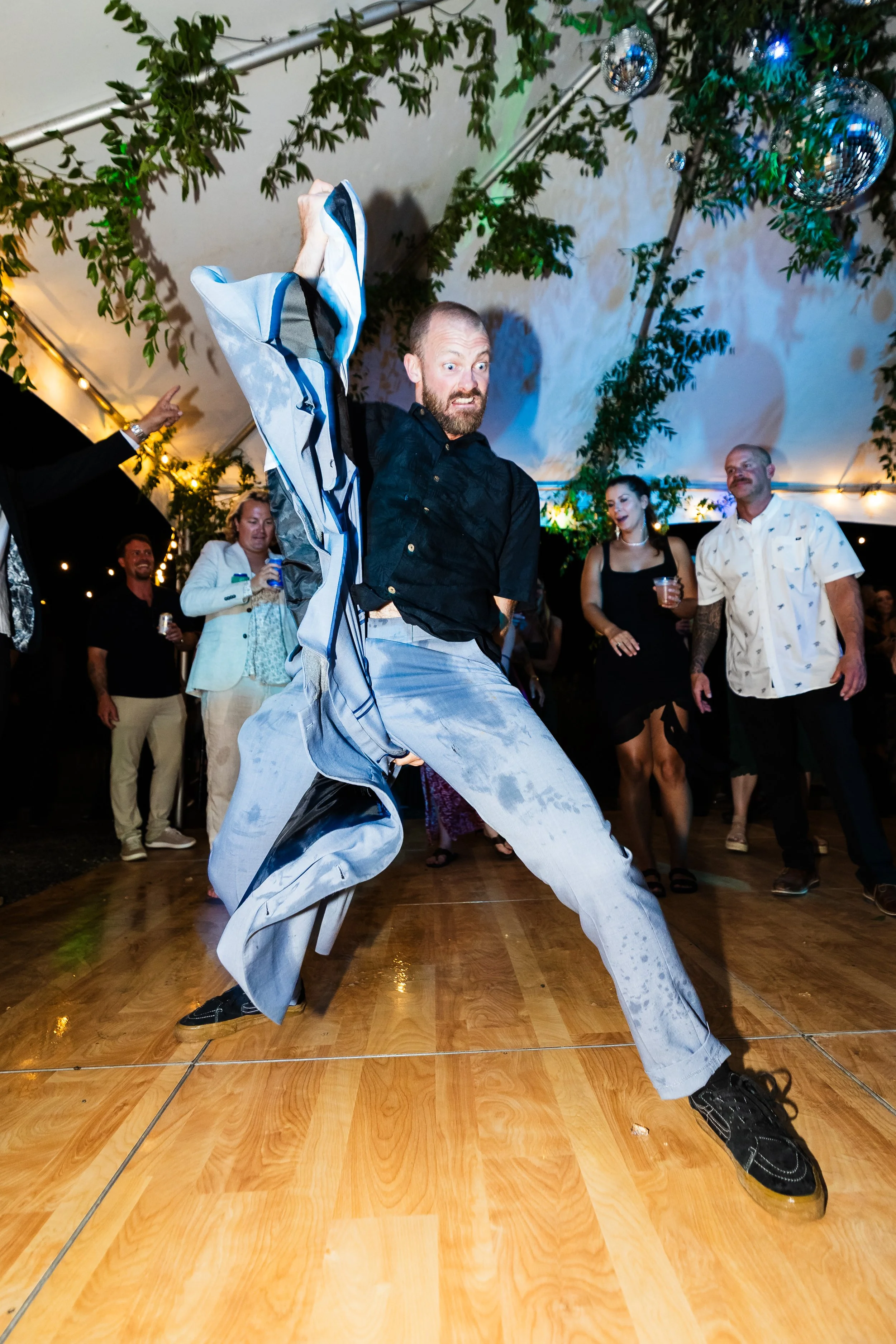 A man with a beard dancing energetically on a wooden floor at a party, wearing a black shirt and light-colored, tie-dye pants, with people watching and celebrating around him under a decorated tent with hanging disco balls and greenery.