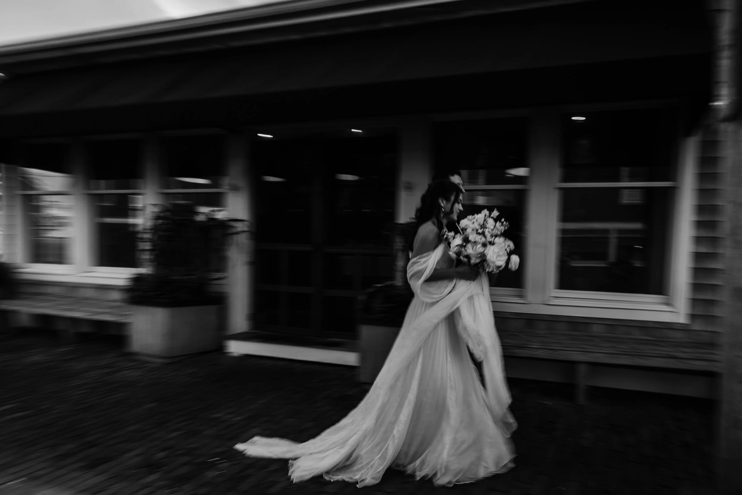 A woman in a wedding dress holding a bouquet of flowers walking outside a building.