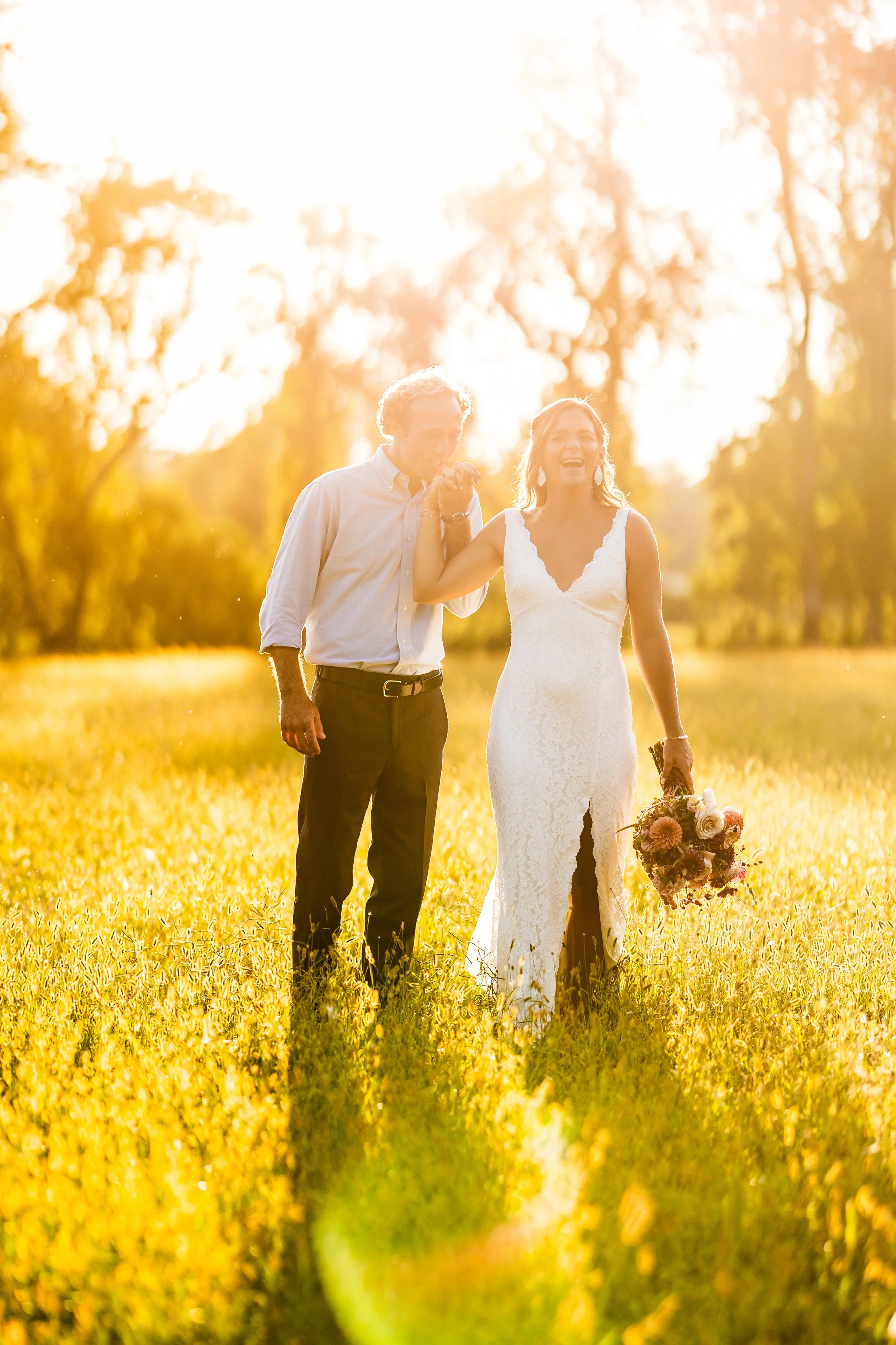 A bride and groom walking in a sunlit field, with the bride holding a bouquet and the groom kissing her hand, during sunset.