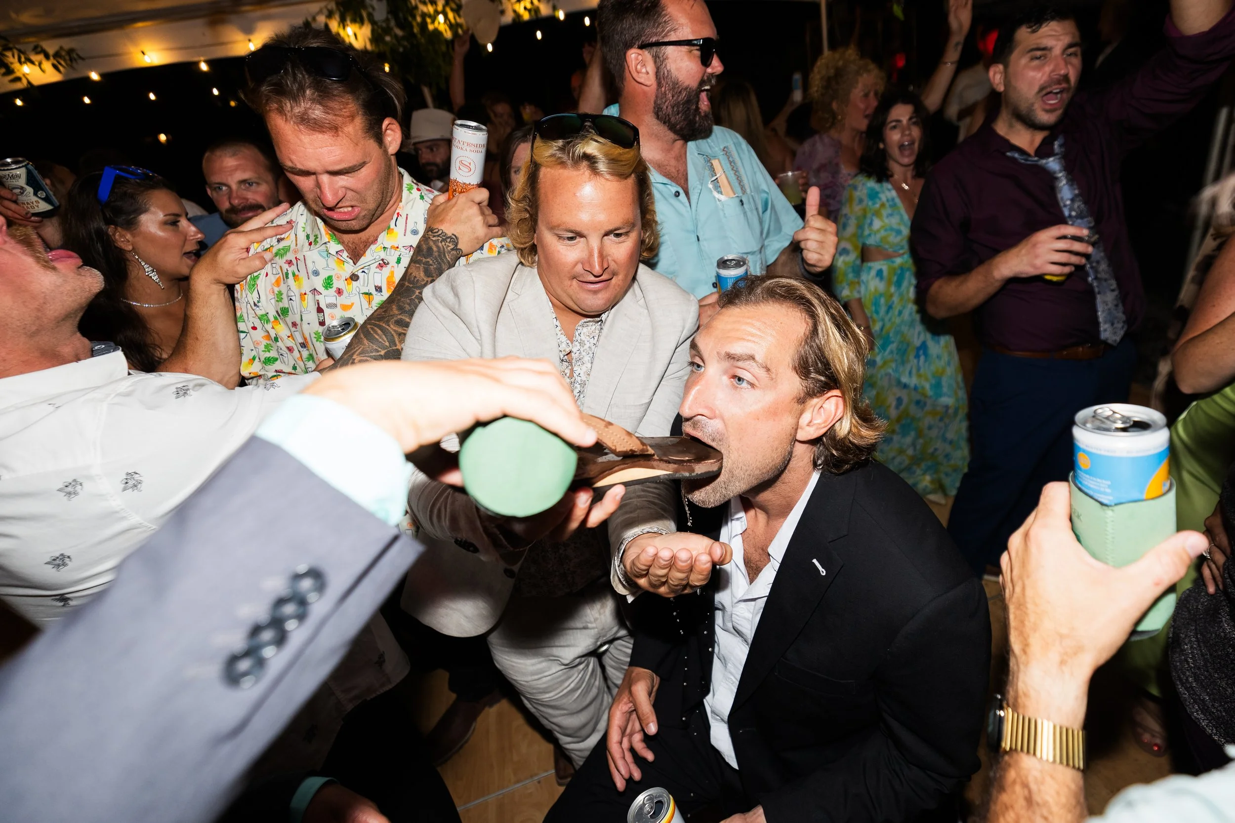 Man in black suit is spitting out a shot of alcohol into his open mouth, held by another person at a lively party with many guests.