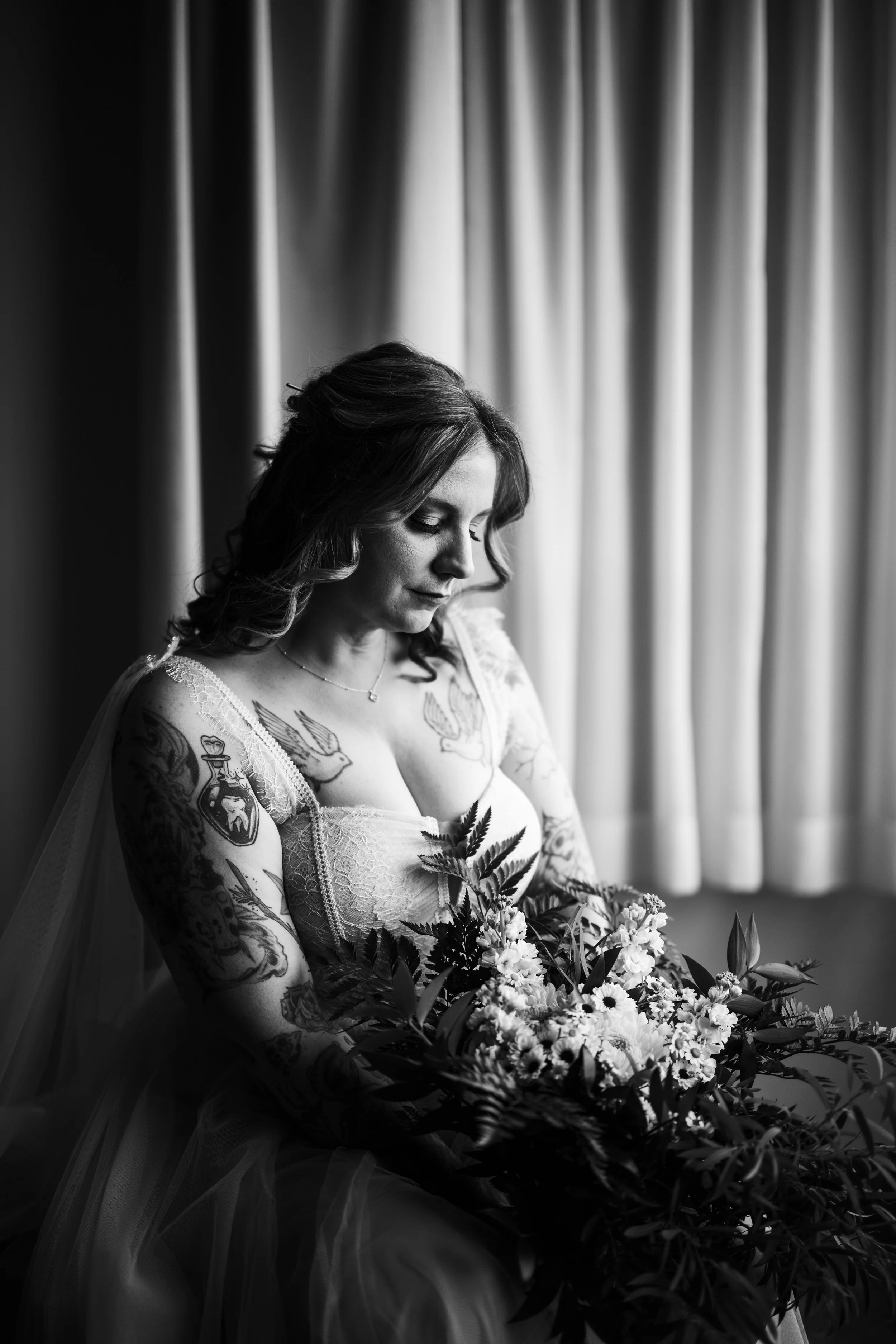 Black and white photo of a woman in a wedding dress sitting by a window with closed curtains, holding a large bouquet of flowers and looking down.