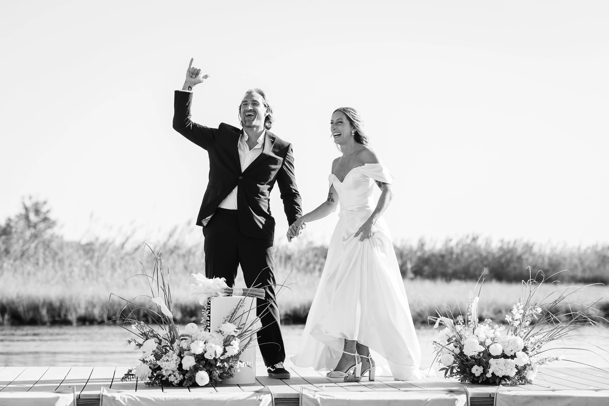 A happy bride and groom on their wedding day, walking hand in hand on an outdoor dock with floral arrangements at their feet, under a clear sky.