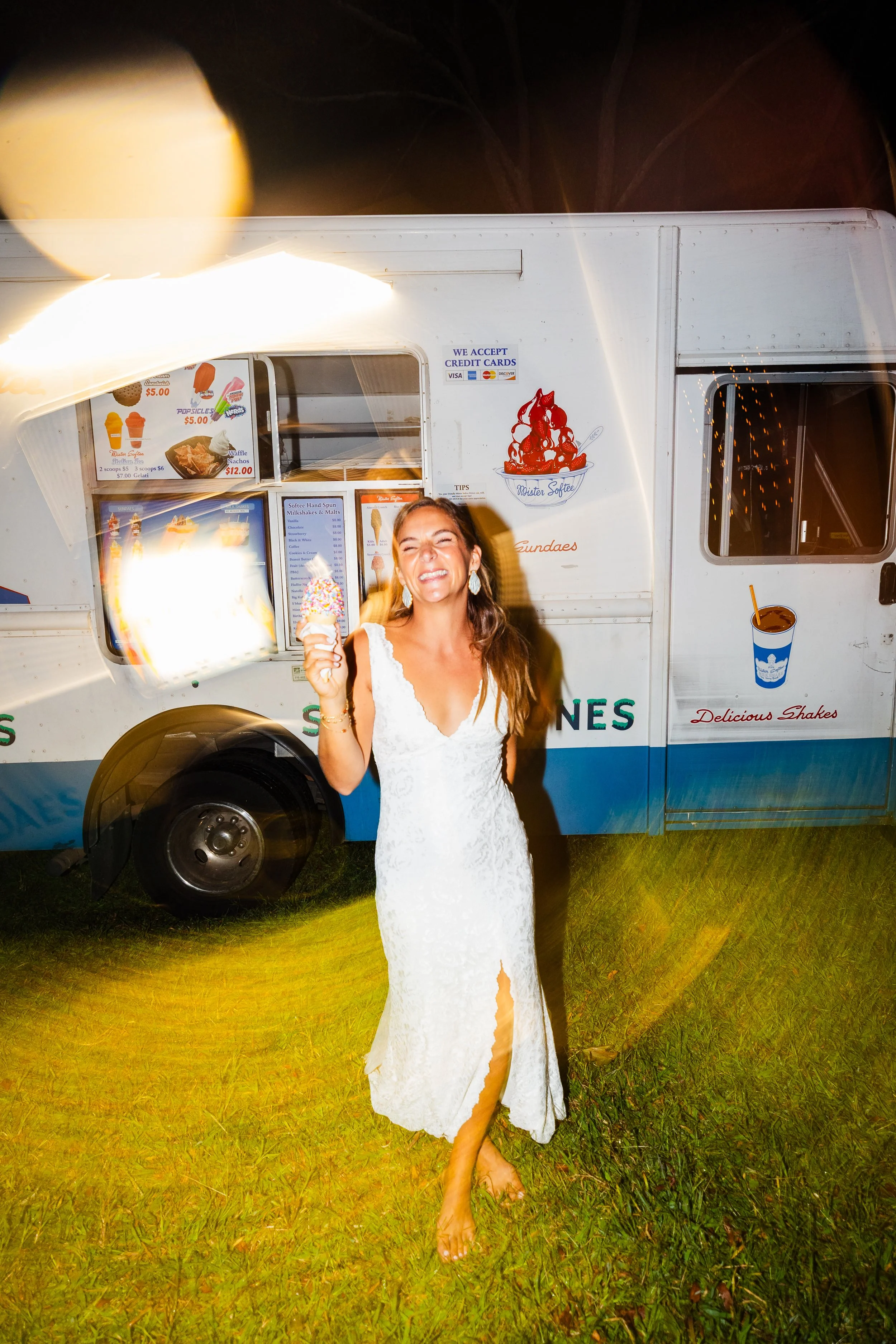 Woman in white lace dress smiling and holding ice cream cone in front of ice cream truck at night.