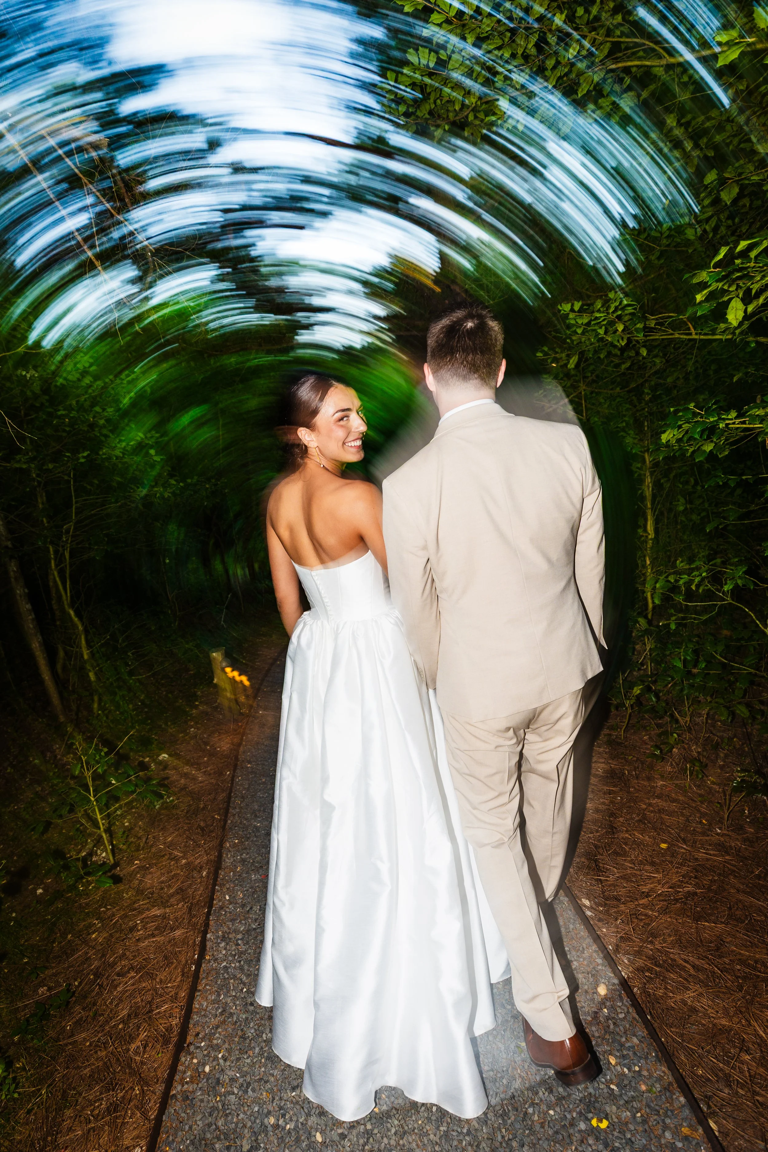 A bride in a white wedding dress and a groom in a beige suit walking together along a wooded path at night, with green and blue swirling lights in the background.