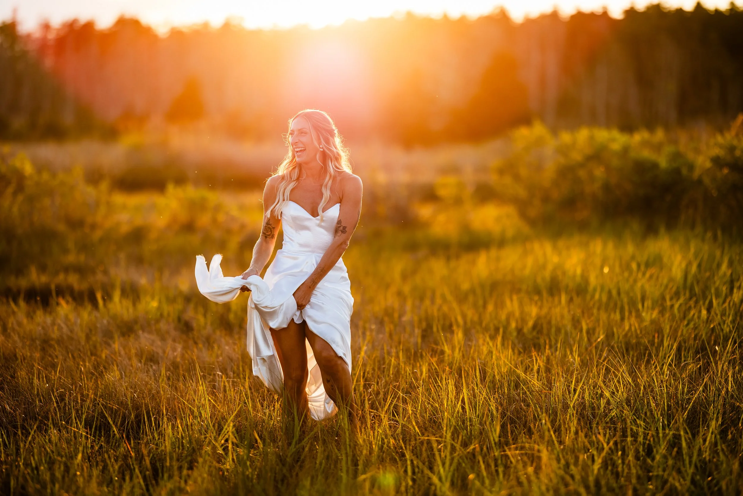 A woman in a white dress running through a grassy field during sunset, smiling and holding her dress.