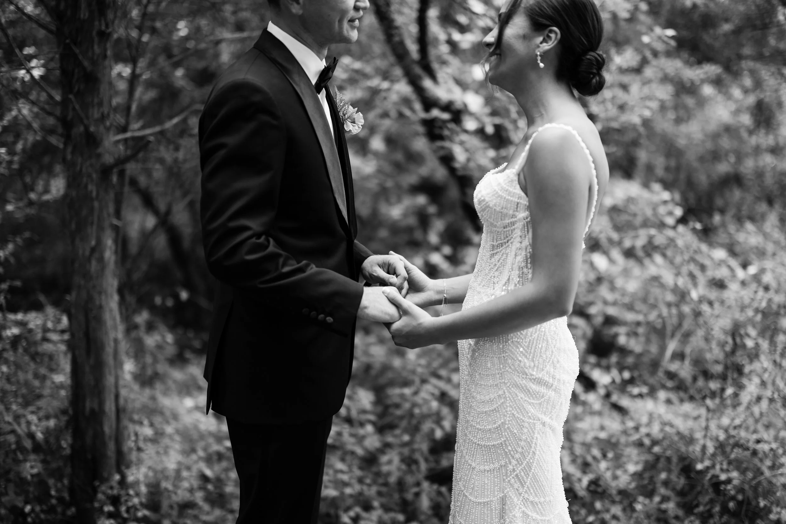 A black and white photo of a bride and groom holding hands and smiling at each other outdoors surrounded by trees.