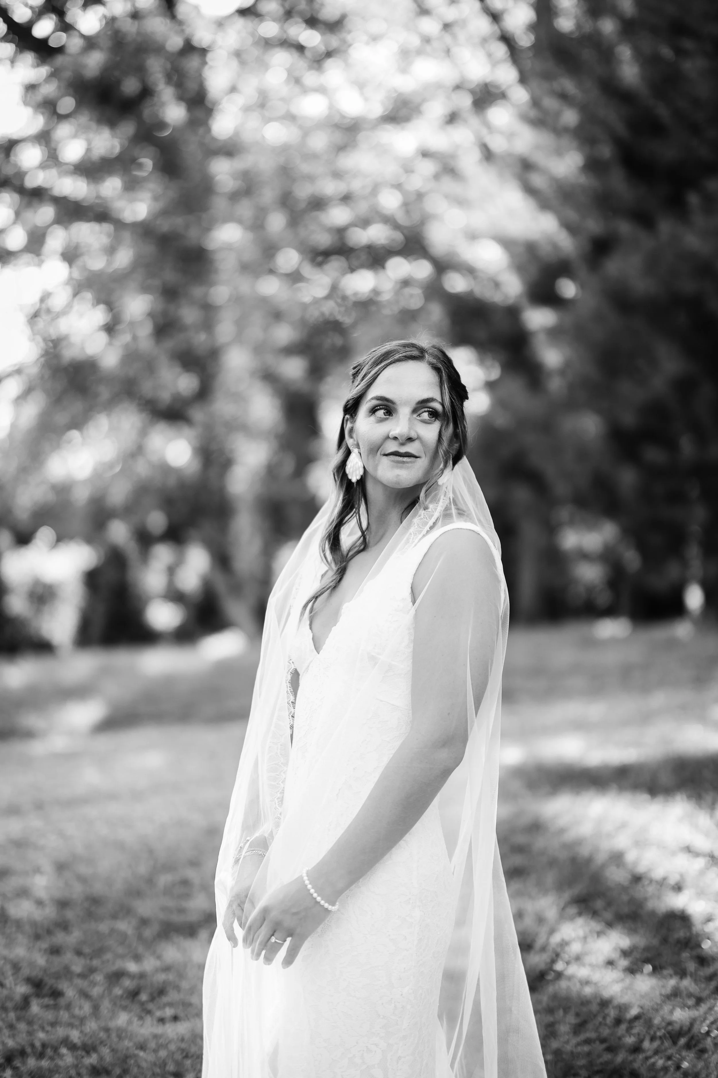 A bride in a wedding dress and veil outdoors, looking to her left with trees and bright sunlight in the background.