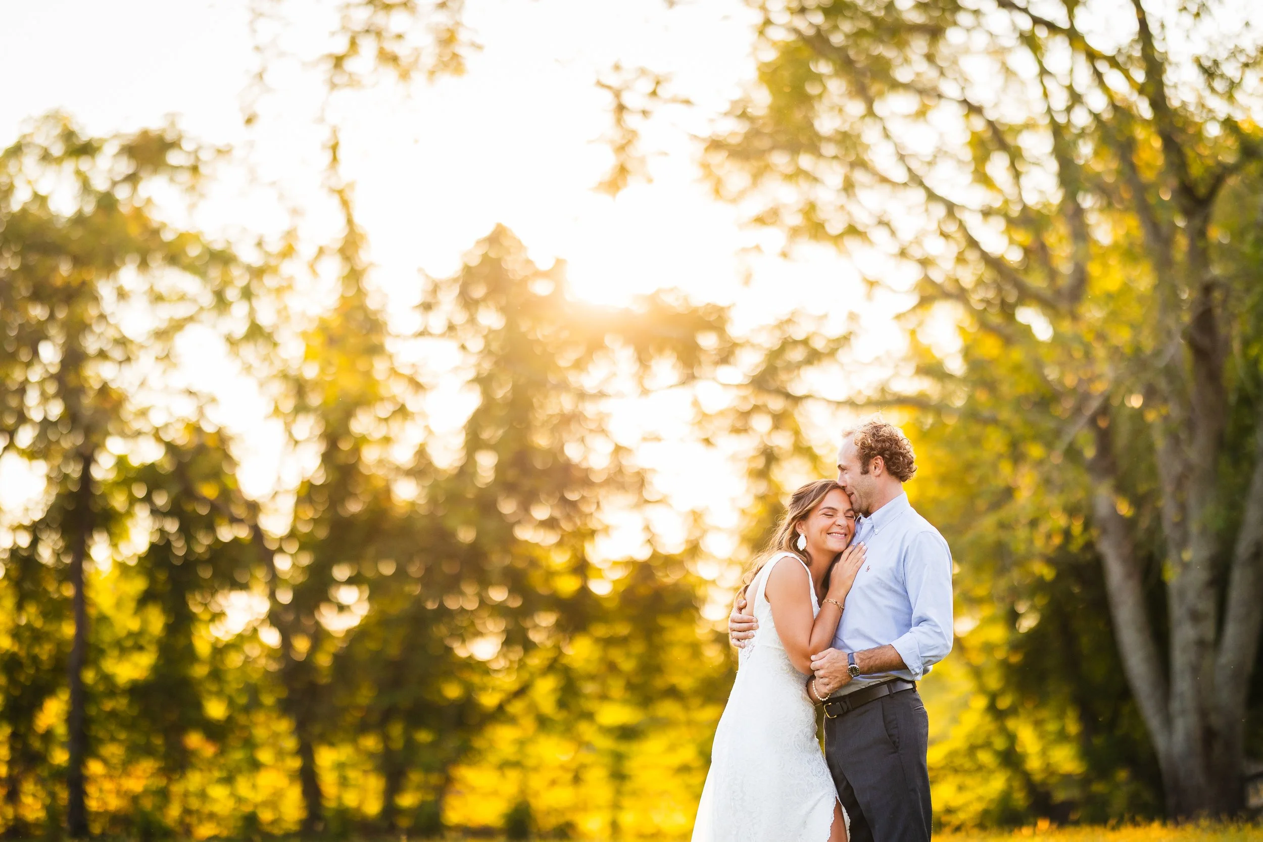 A couple embracing outdoors during sunset, with the woman smiling and the man kissing her forehead, surrounded by trees and warm sunlight.