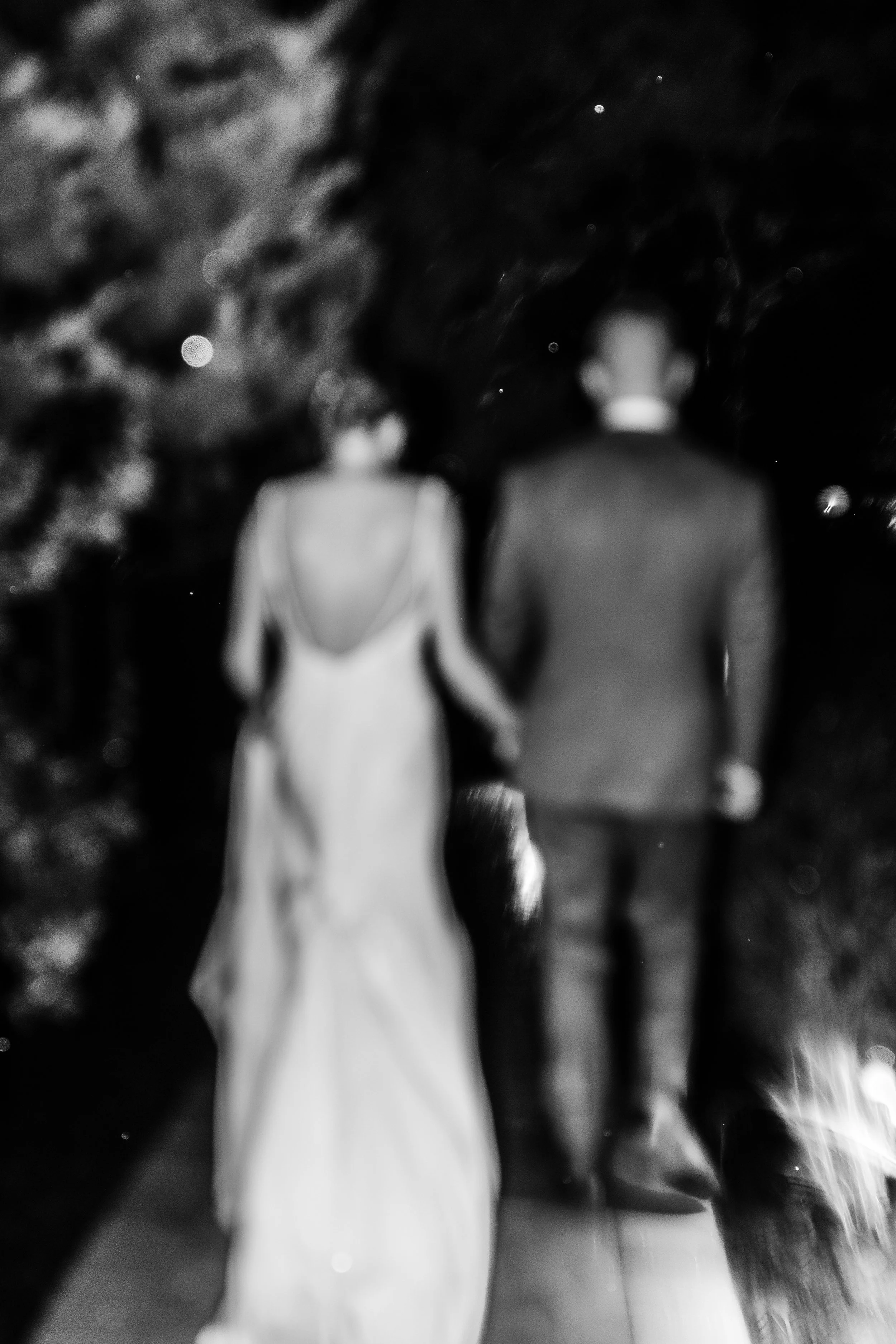 A black and white photograph of a couple walking outdoors at night, holding hands, with the woman wearing a long dress and the man in a suit.