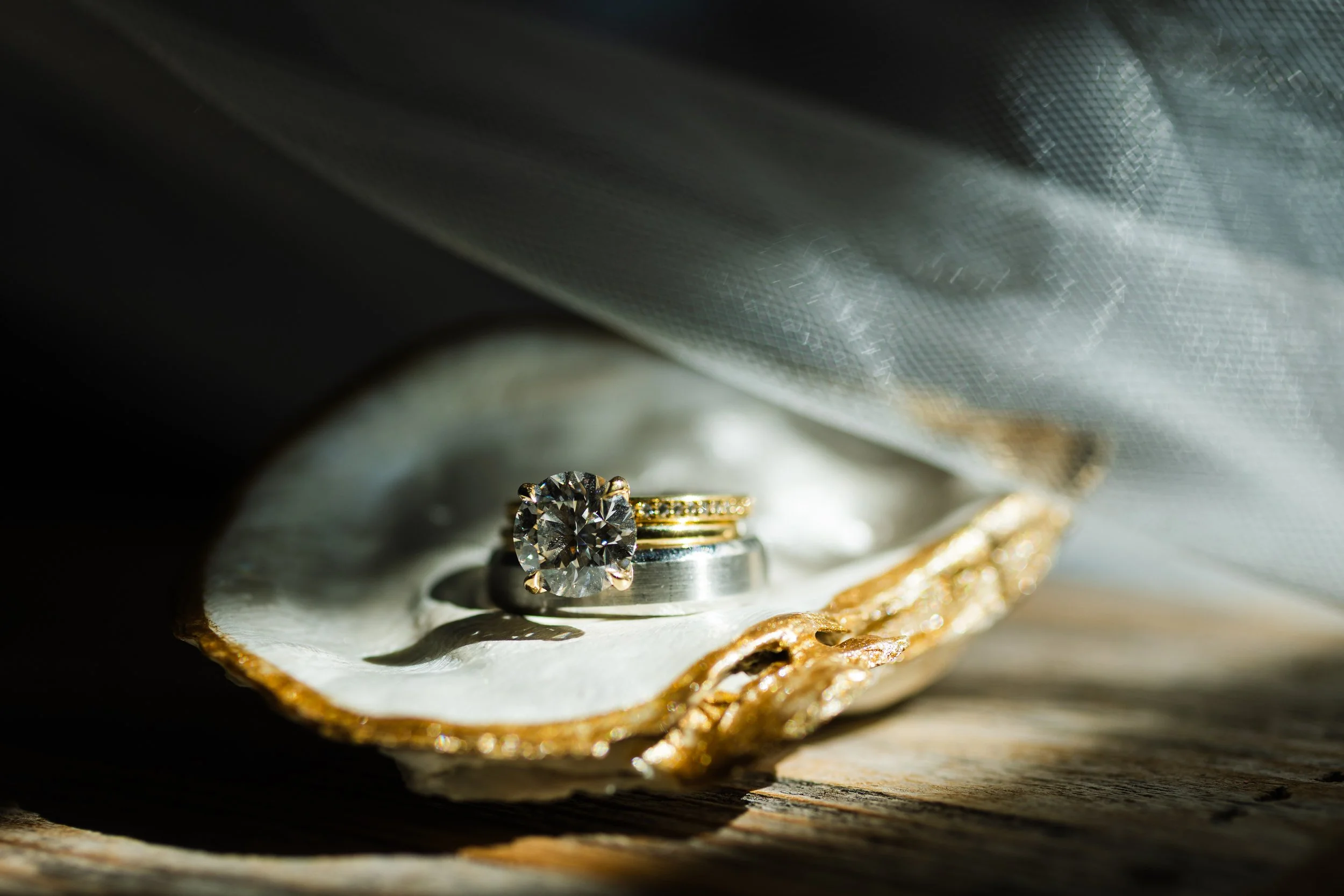Close-up of a diamond engagement ring with a large stone, resting inside a clam shell with gold decorative edges, on a wooden surface.