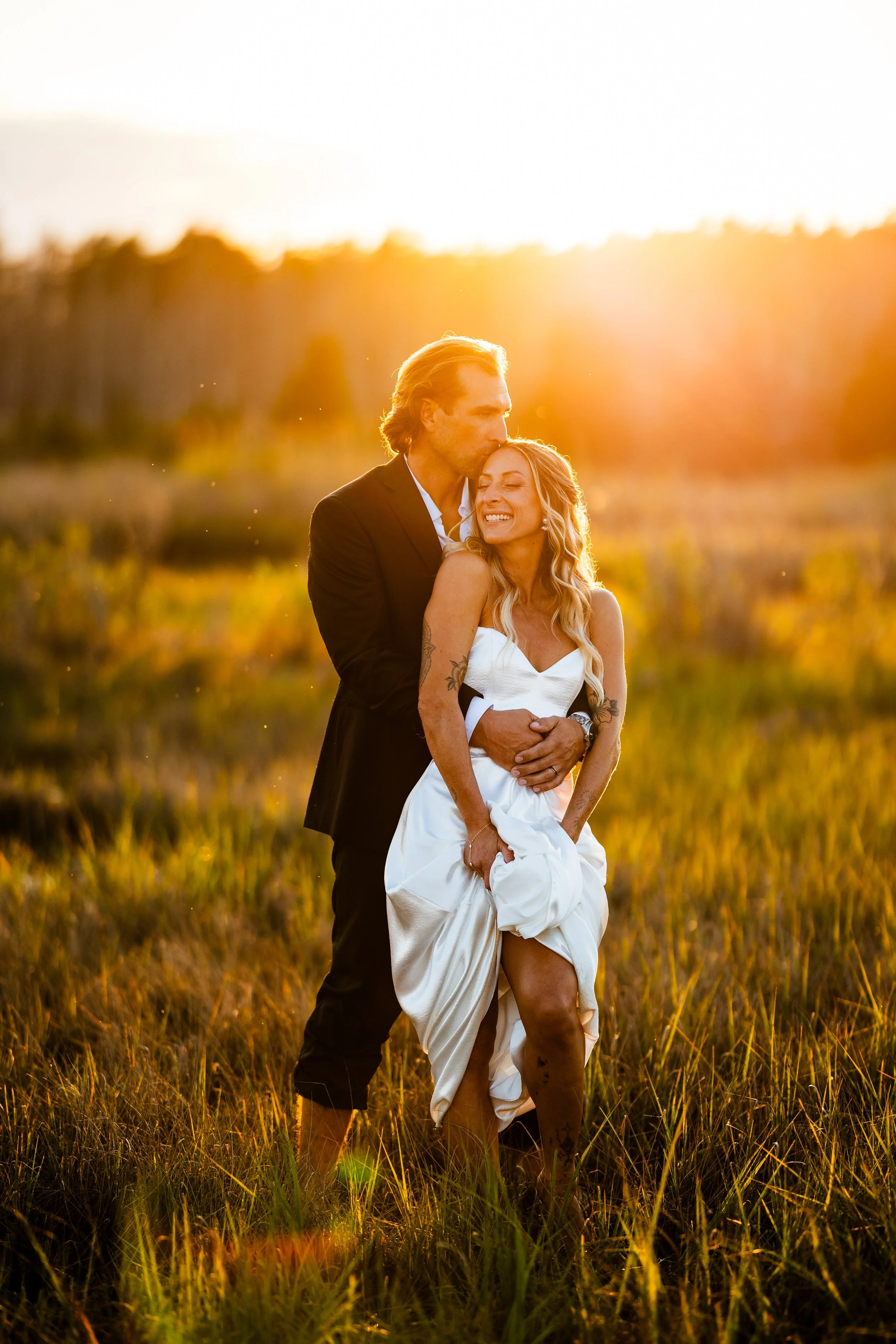 A couple standing in a field at sunset, with the man kissing the woman on the head and holding her, both smiling.