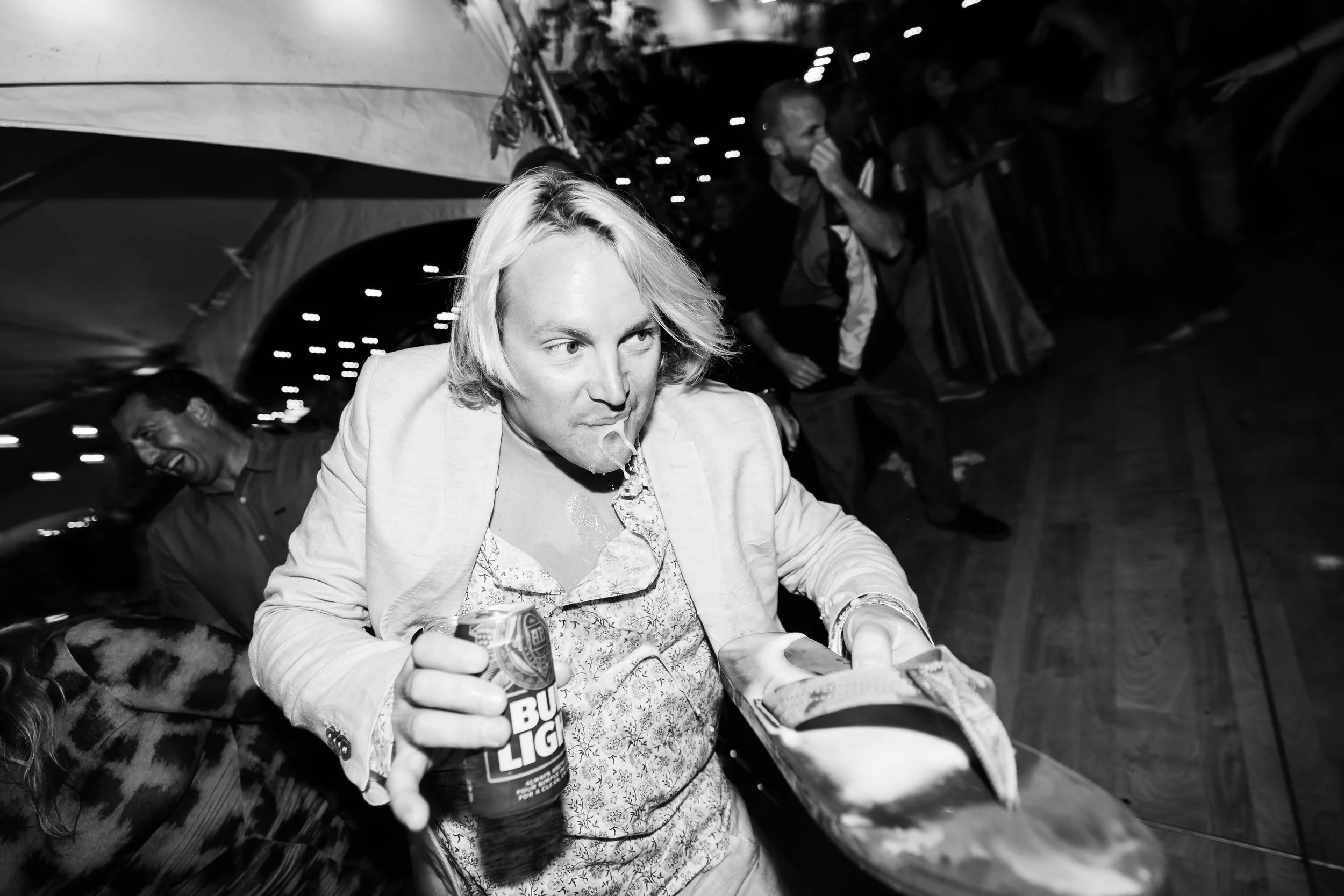 A woman with blond hair is holding a Bud Light beer can and a pizza on a round tray, leaning over a bar counter at a lively indoor event.