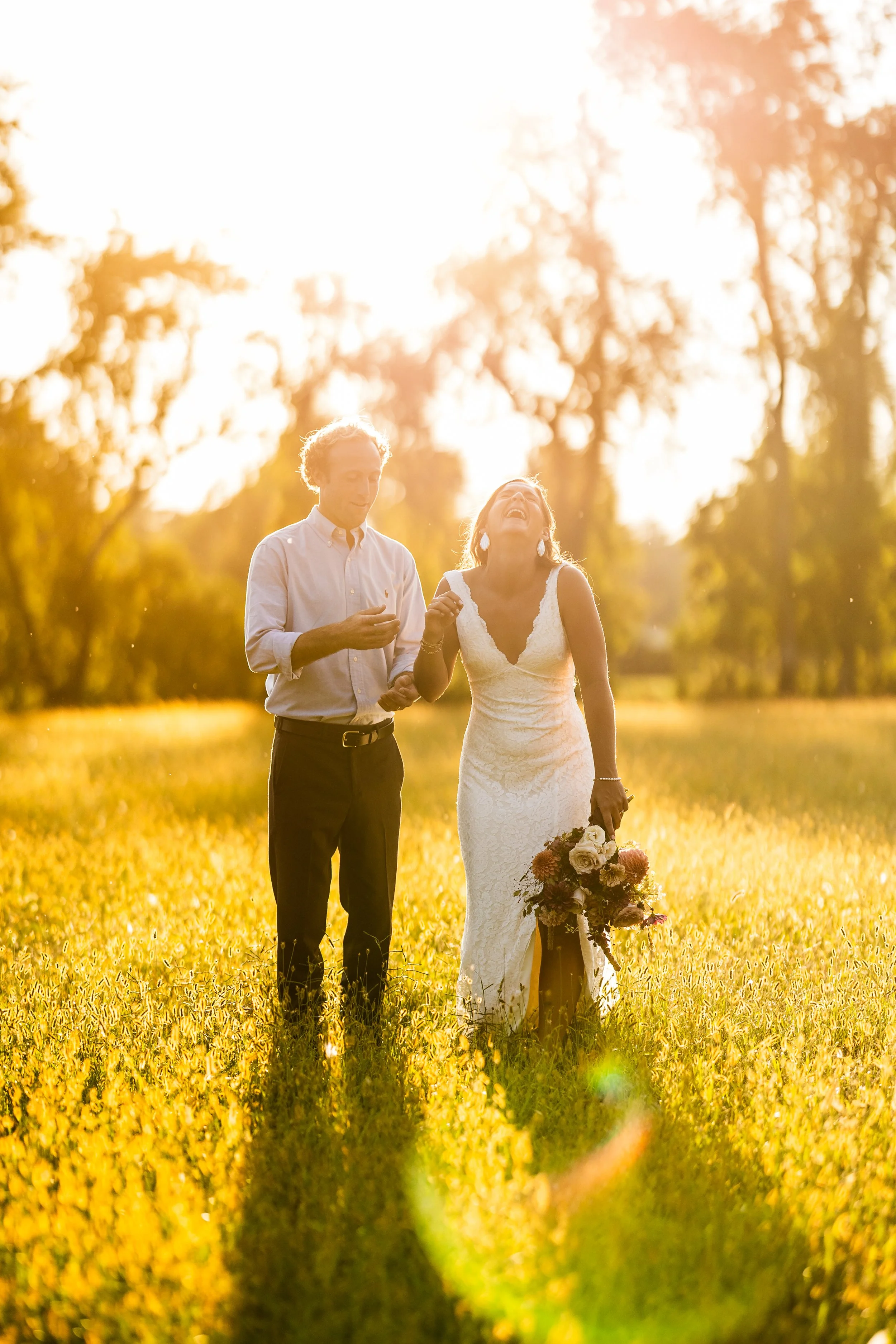 A bride and groom walking through a field of yellow flowers during sunset, smiling and laughing, with trees in the background.