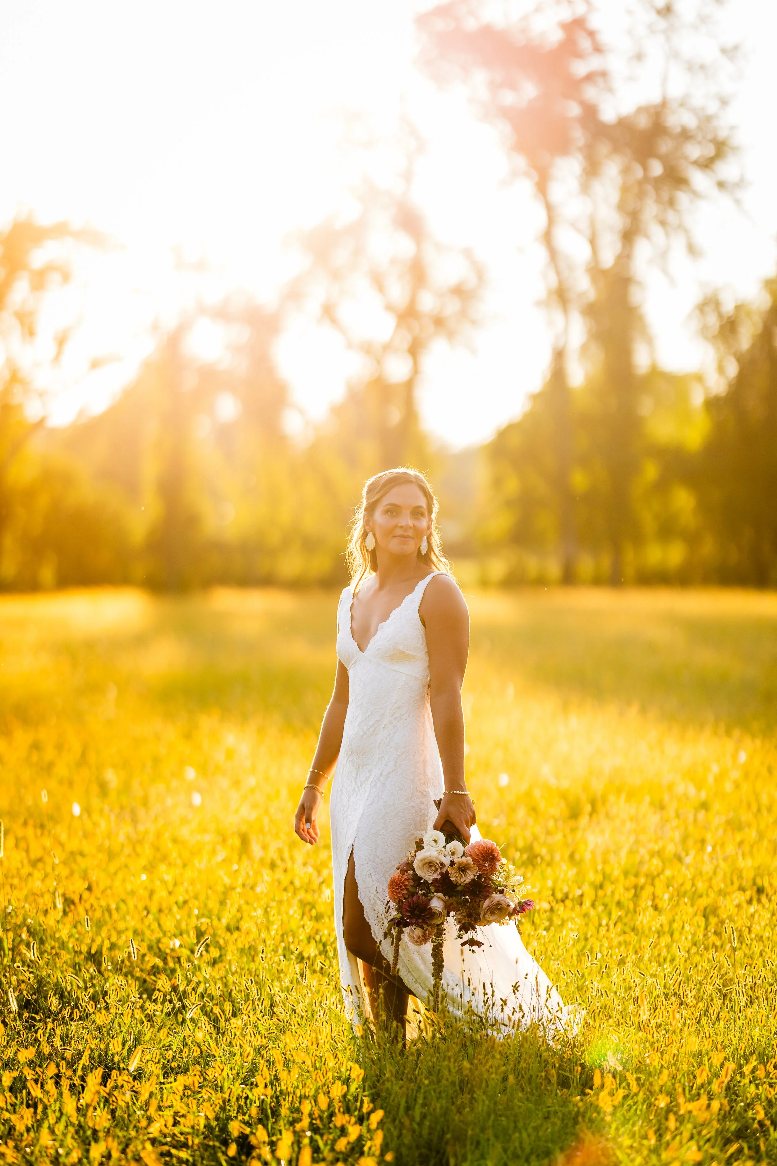 A woman in a white wedding dress holding a bouquet of flowers walking in a sunlit field of yellow flowers.