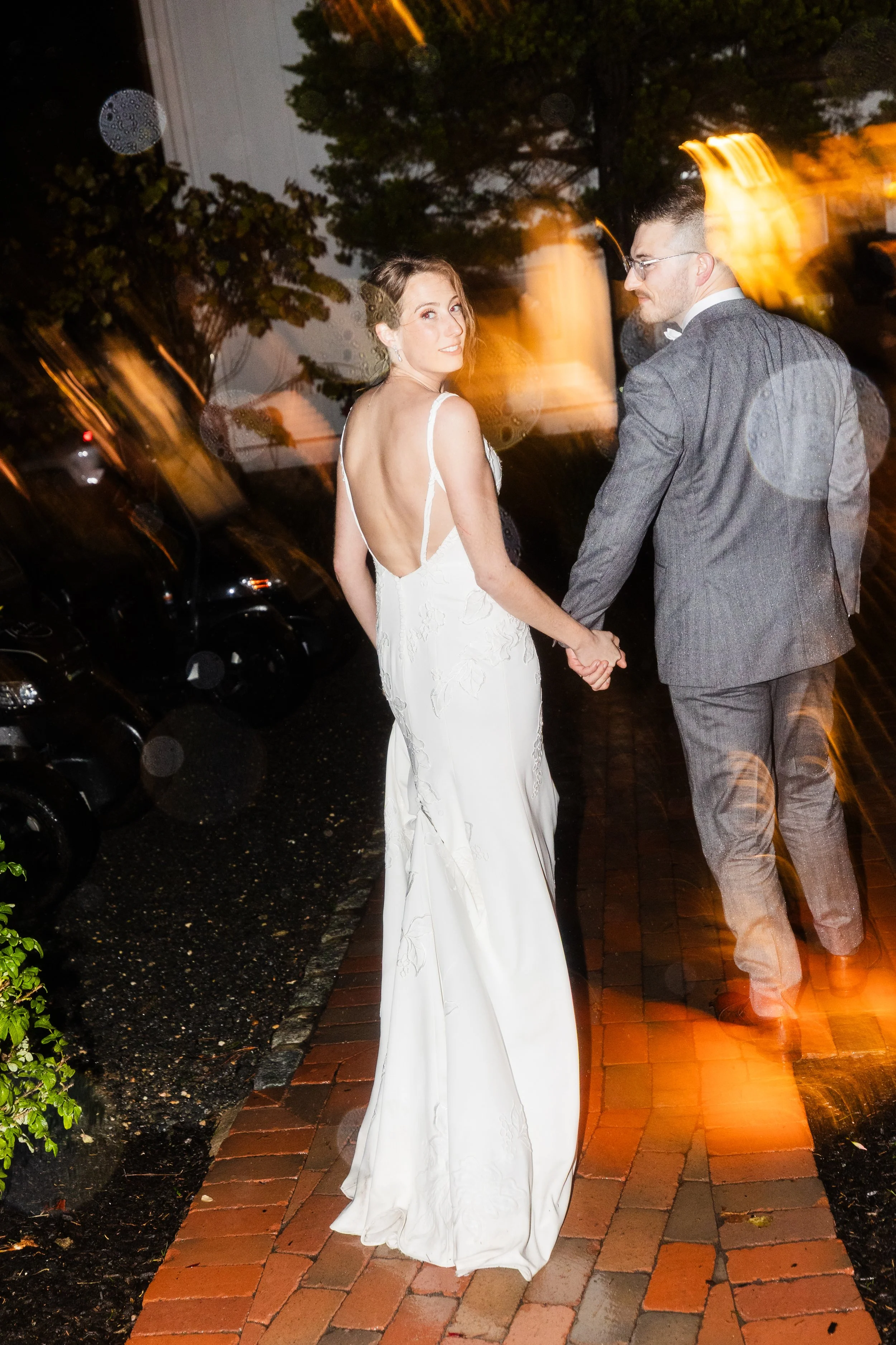 A bride and groom holding hands and walking on a brick sidewalk at night, with trees and parked cars in the background, and a motion blur effect from rain or movement.