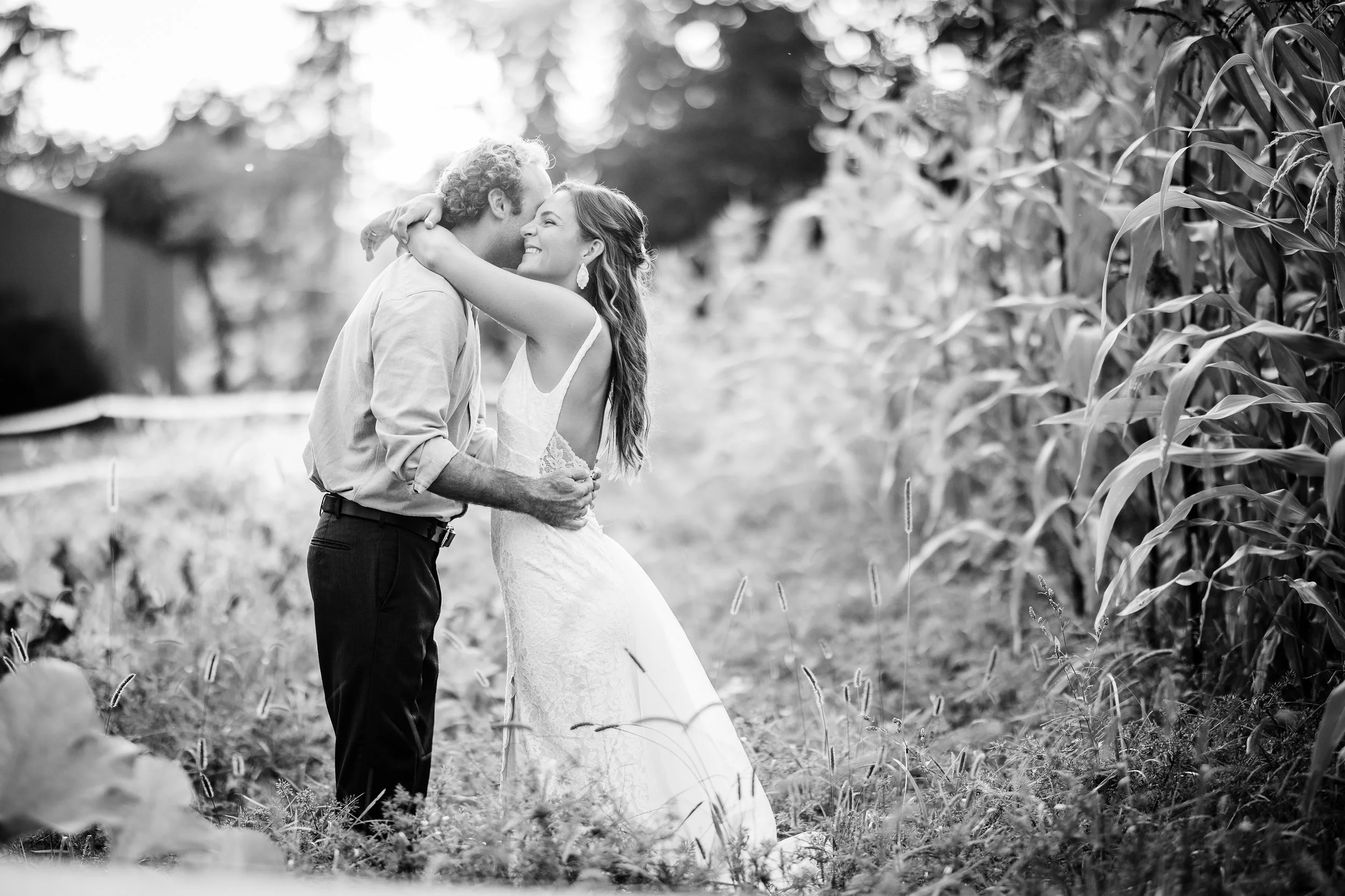 A black-and-white photo of a couple embracing in a field of tall corn plants. The woman is wearing a sleeveless lace dress and the man is in a light-colored shirt and dark pants. They are smiling and looking at each other lovingly.