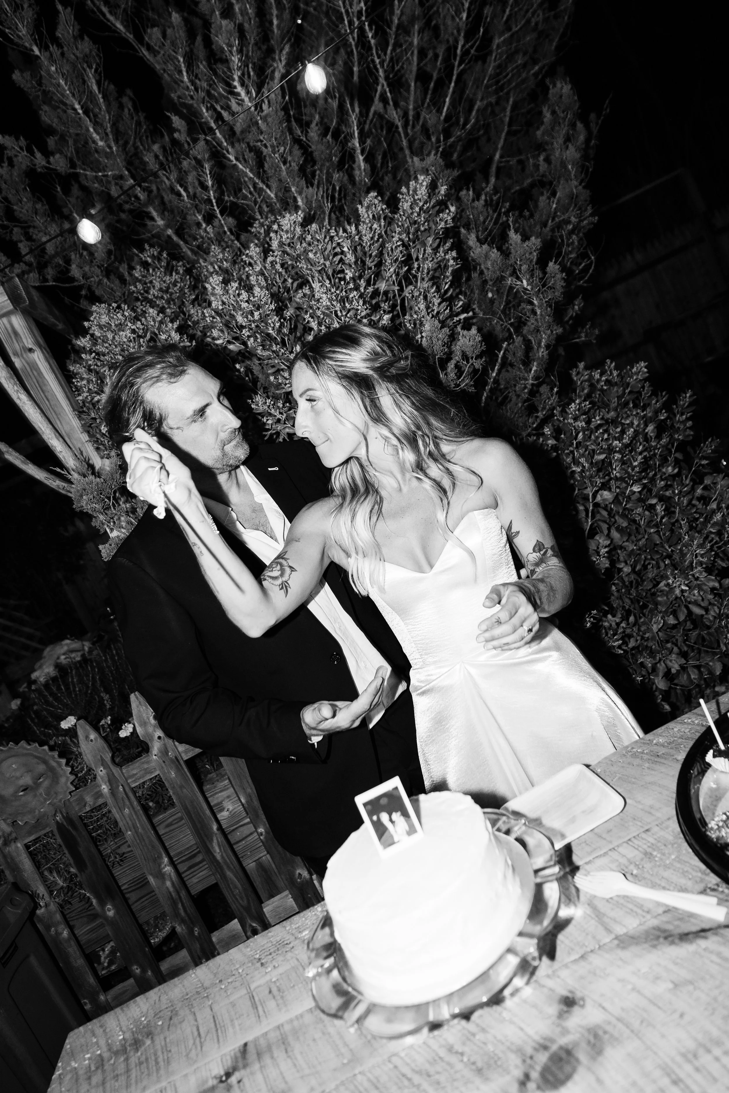 Black and white photo of a couple dancing closely at their wedding reception, with a wedding cake on the table in front of them, outdoors at night with string lights overhead.