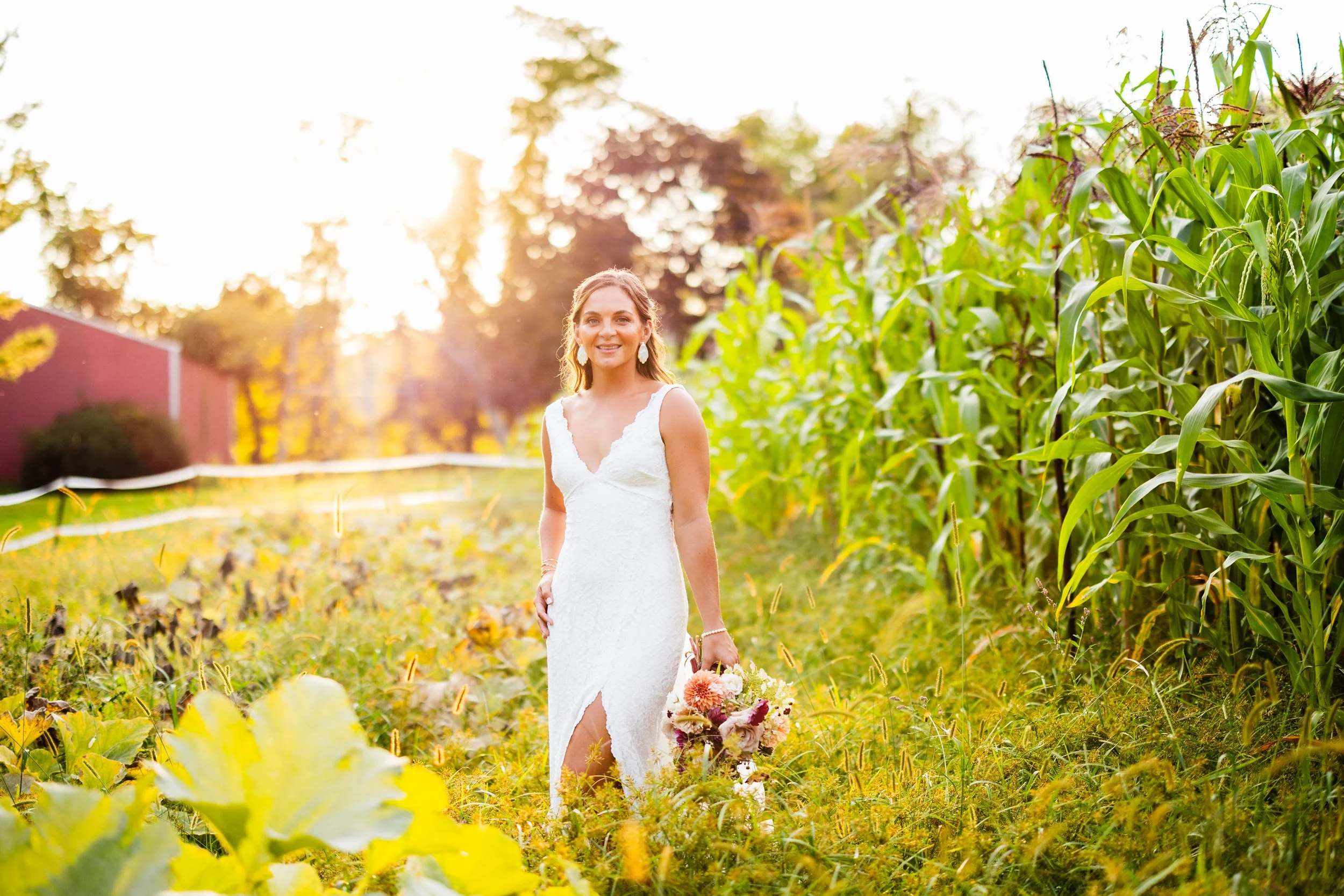 A woman in a white wedding dress holding a bouquet, walking through a field of tall grass and crops at sunset.