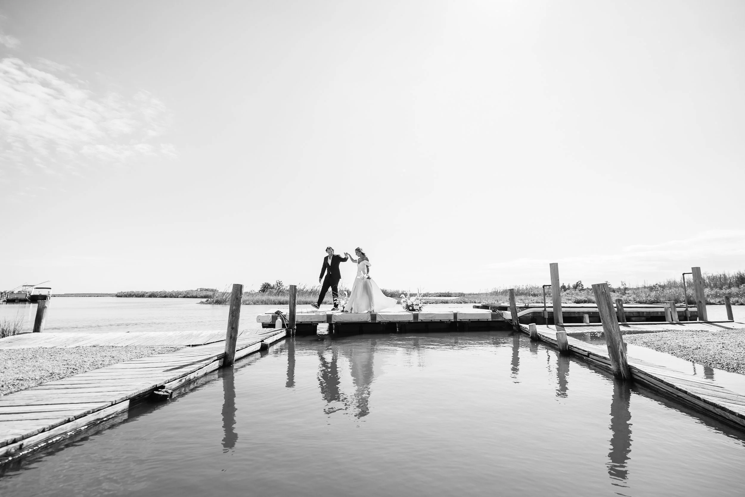 A bride and groom holding hands on a dock over water, with boats in the distance under a clear sky, viewed in black and white.