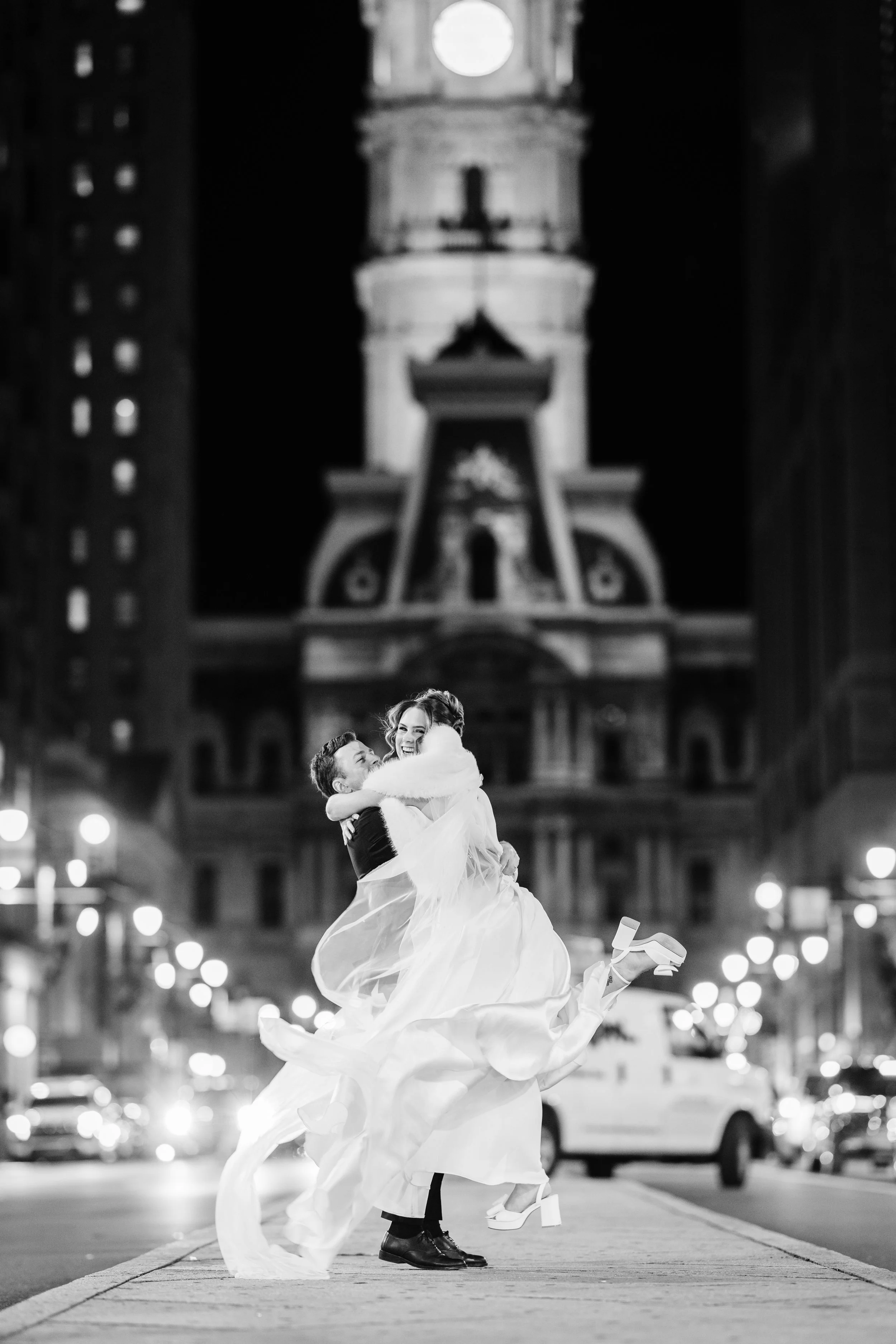 A couple in wedding attire dancing and embracing on a city street at night, with a prominent clock tower in the background and street lights illuminating the scene.