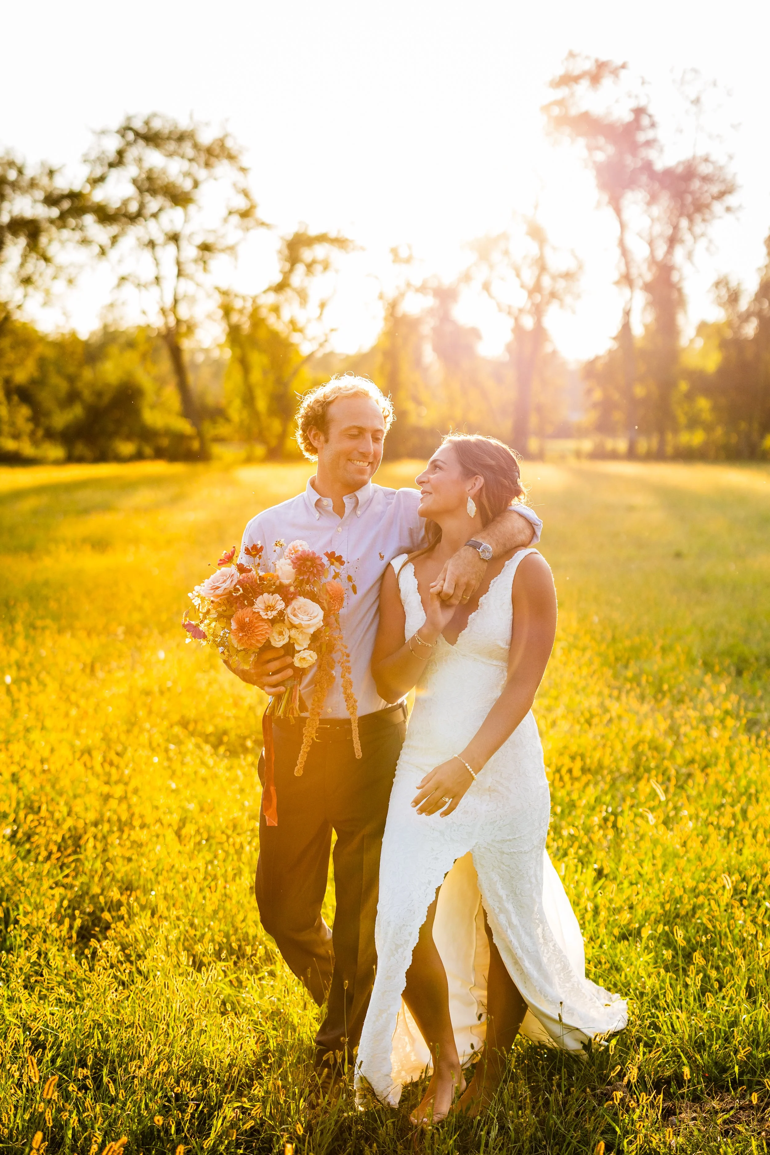 A newlywed couple walking together in a sunlit grassy field during sunset, with the groom holding a bouquet of flowers and the bride smiling at him.