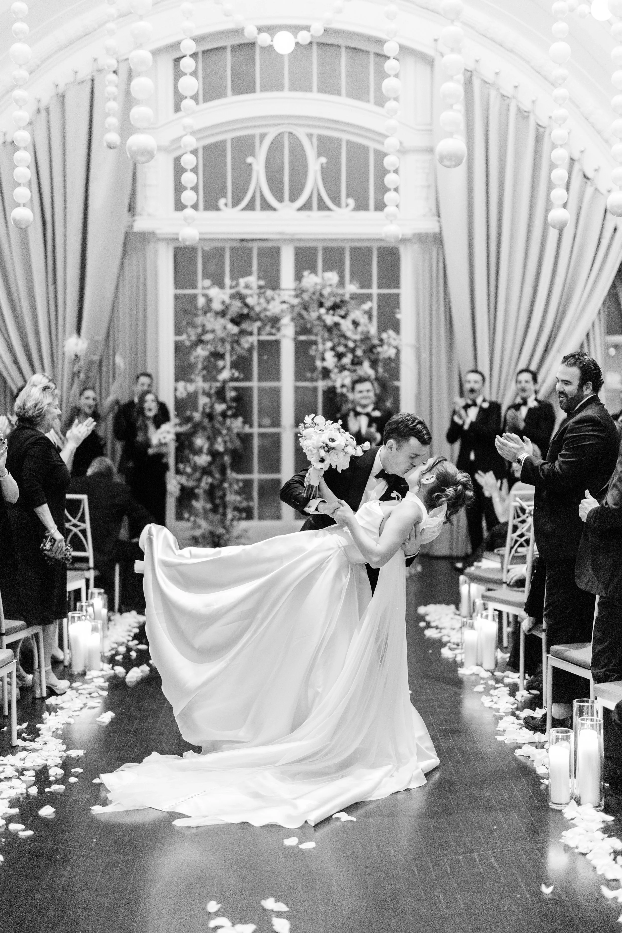 A bride and groom sharing a dance at their wedding reception, with wedding guests clapping and celebrating in a decorated indoor venue.