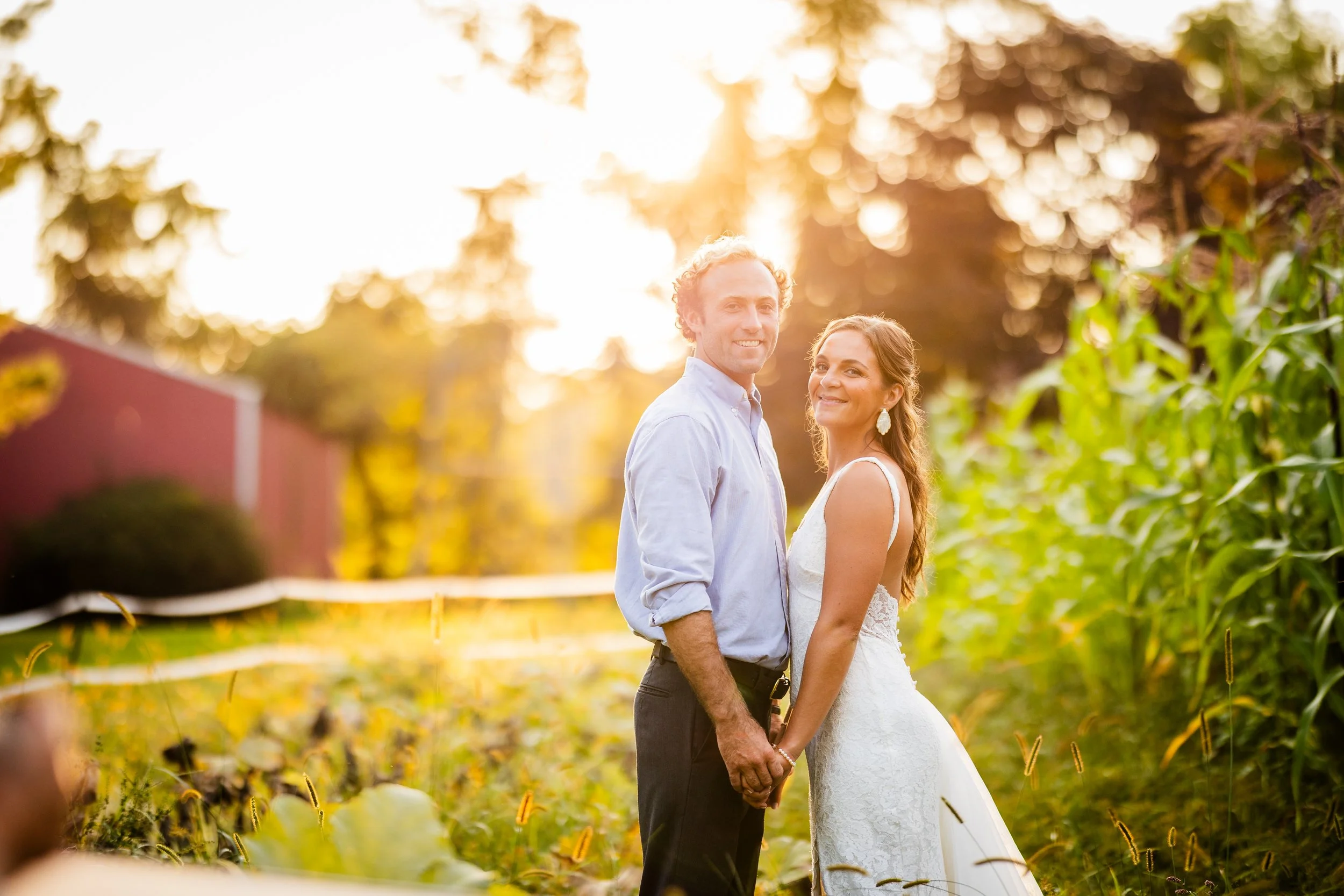 A newlywed couple holding hands and smiling outdoors at sunset, surrounded by lush green plants and trees.