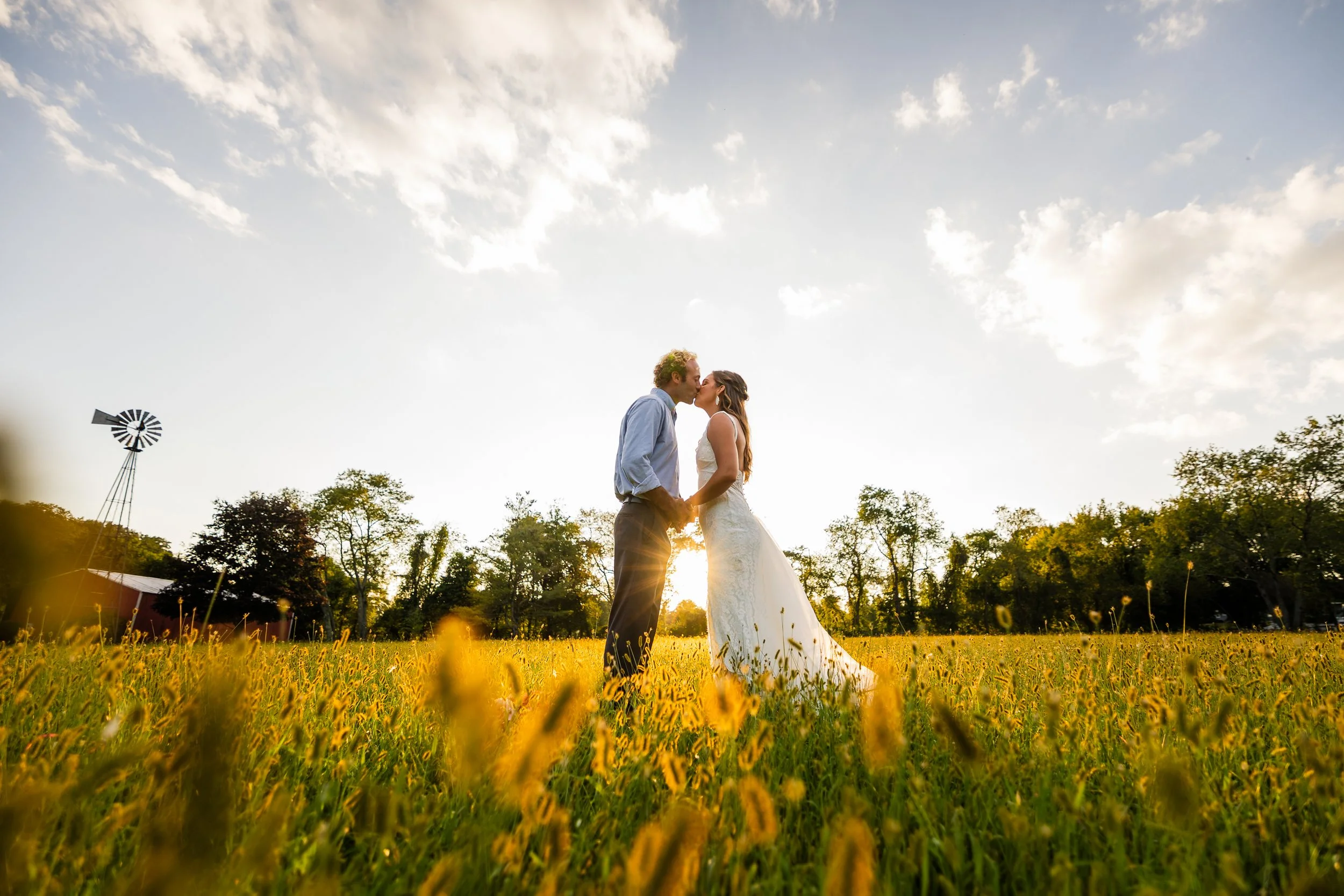 A couple dressed in wedding attire standing in a field of yellow flowers at sunset, kissing with trees and a windmill in the background.