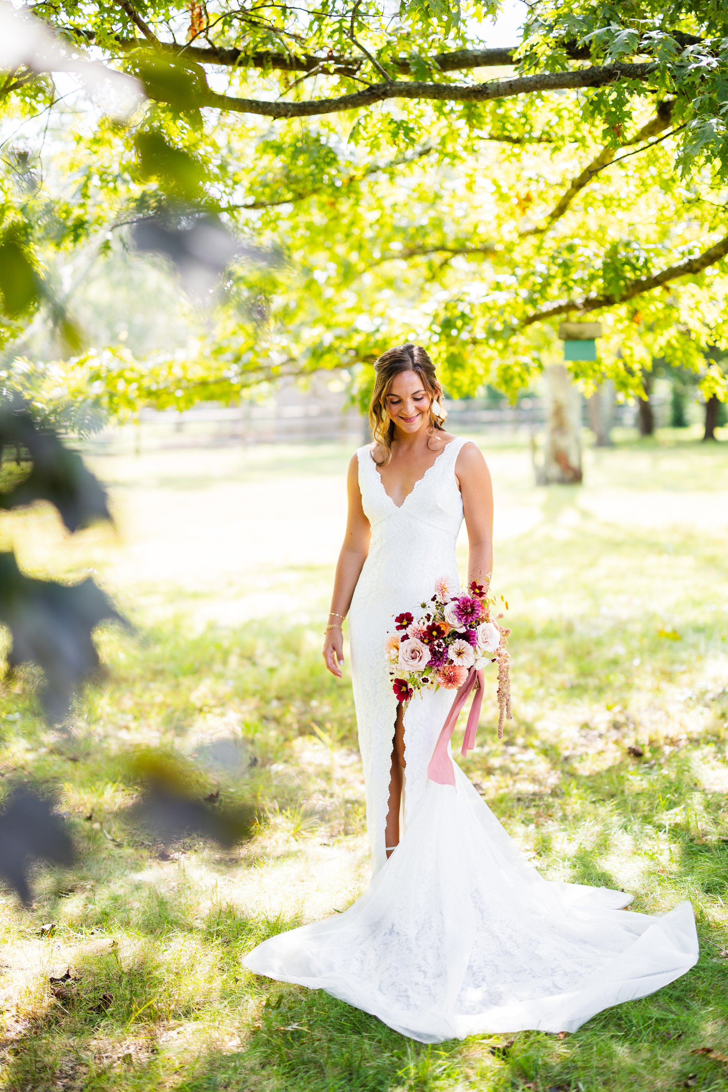 A woman in a white wedding dress holding a bouquet of pink, purple, and white flowers, standing outdoors under a tree with green leaves, sunlight filtering through.