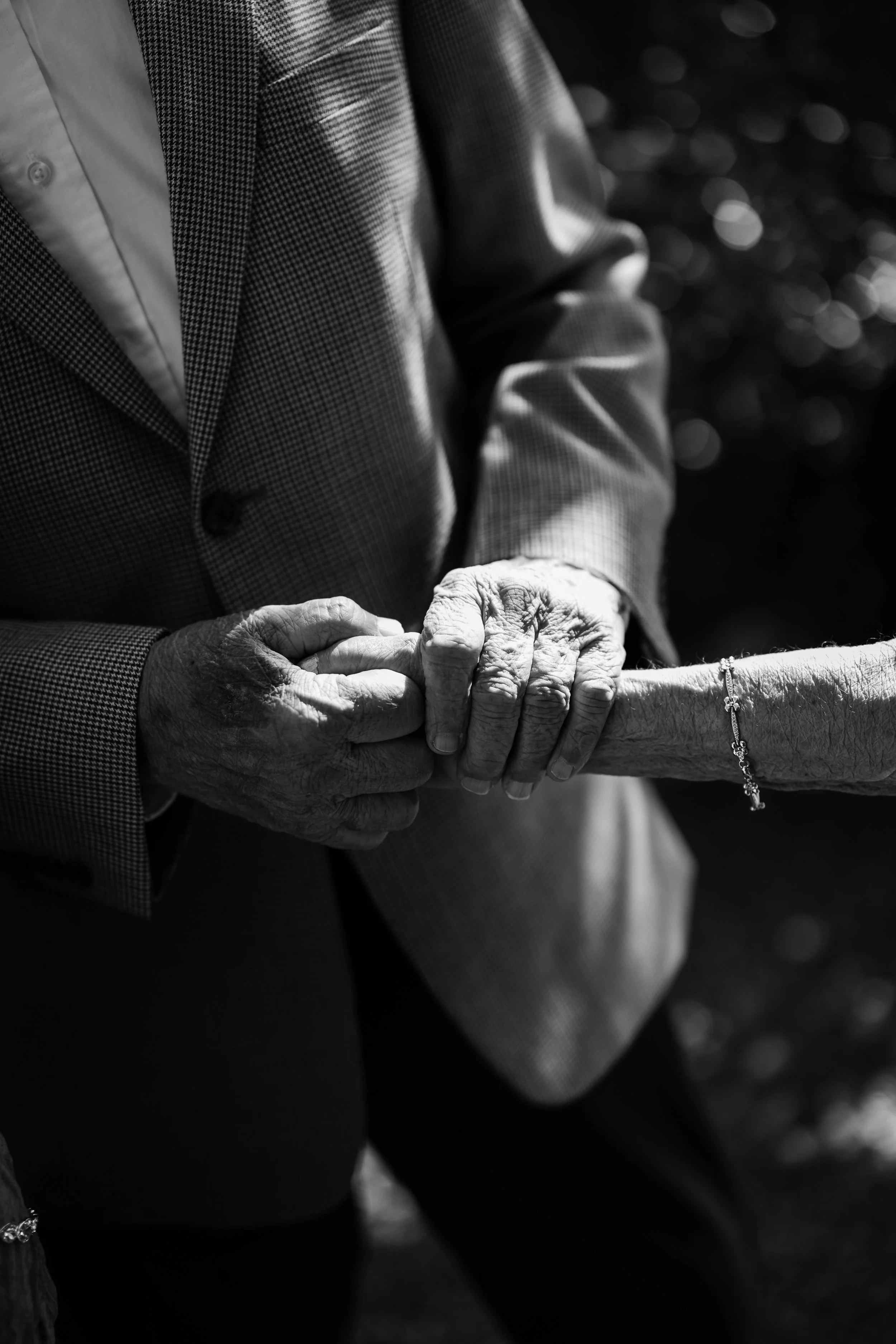 Close-up of an elderly person's hands being gently held by another person, with both wearing formal clothing, in black and white.