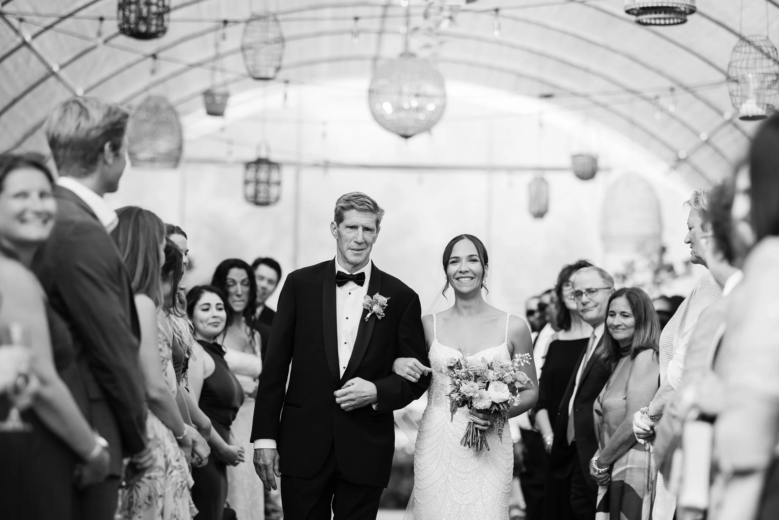 A bride walking down the aisle with her father during a wedding ceremony, surrounded by guests in a decorated outdoor venue.