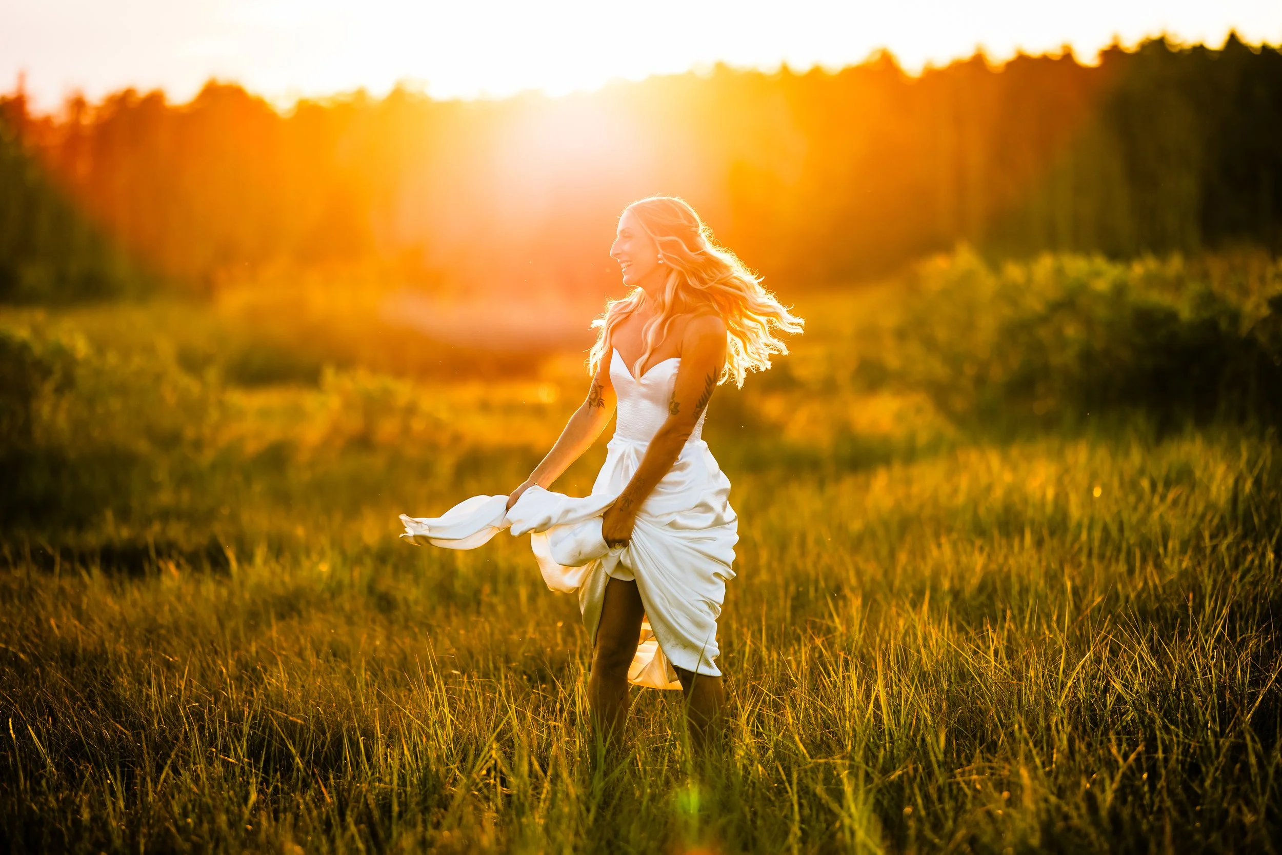 A woman in a white dress is standing in a grassy field during sunset, smiling and holding her dress as her hair blows in the wind.