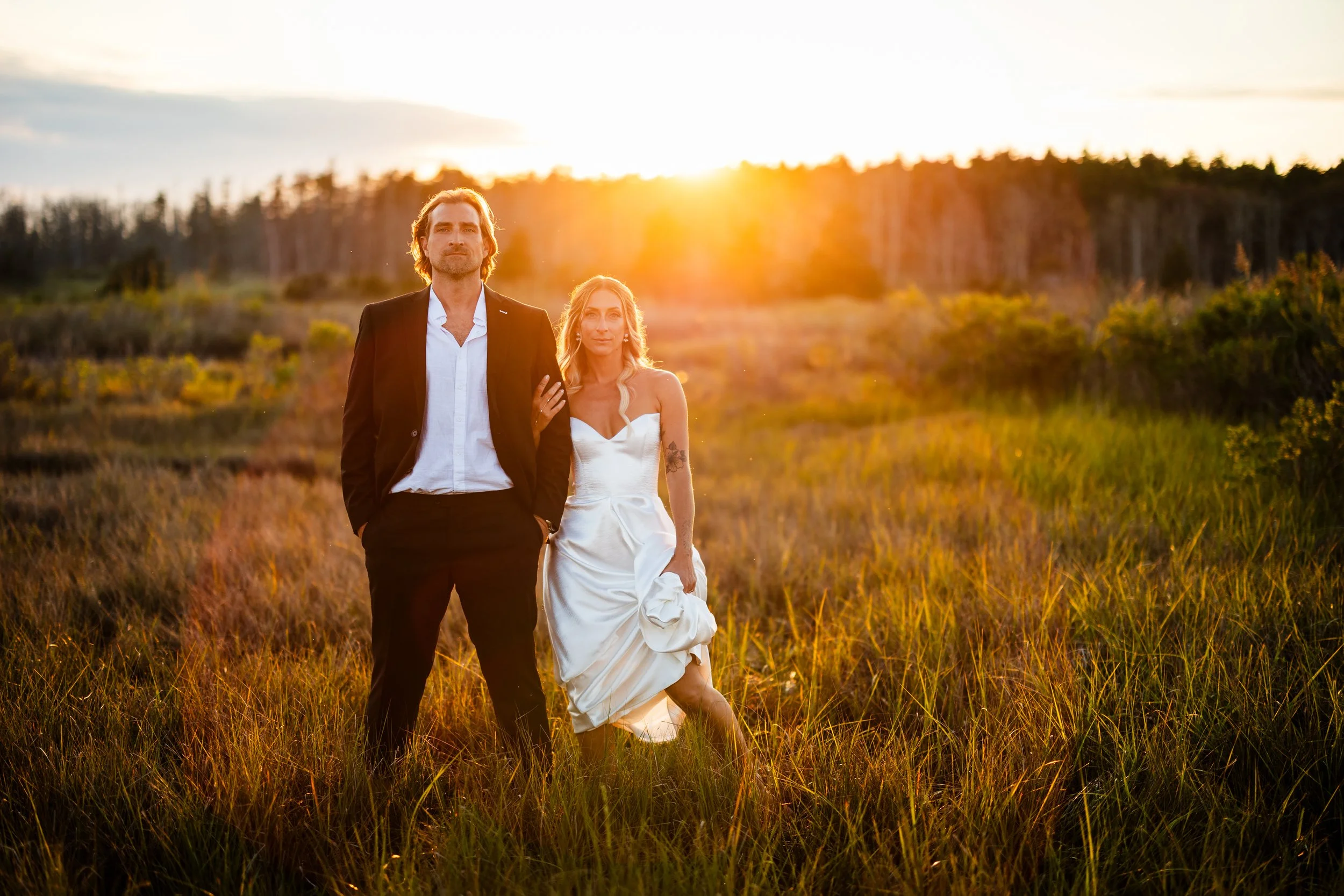 A man in a black suit and white shirt, and a woman in a white dress, standing together in a grassy field at sunset with trees in the background.