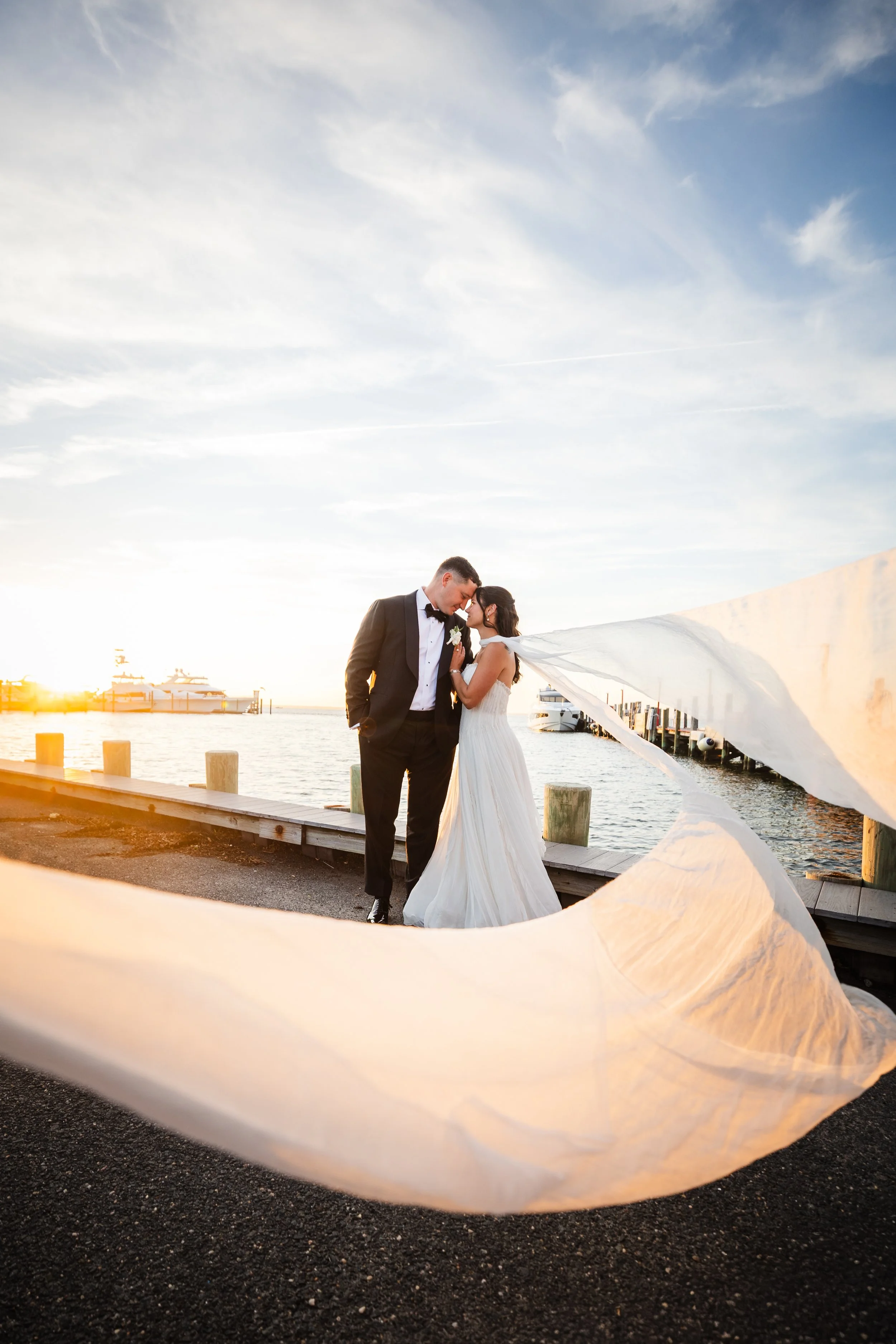 A bride and groom on a dock by the water at sunset, standing close and touching foreheads, with a flowing white veil in the foreground.