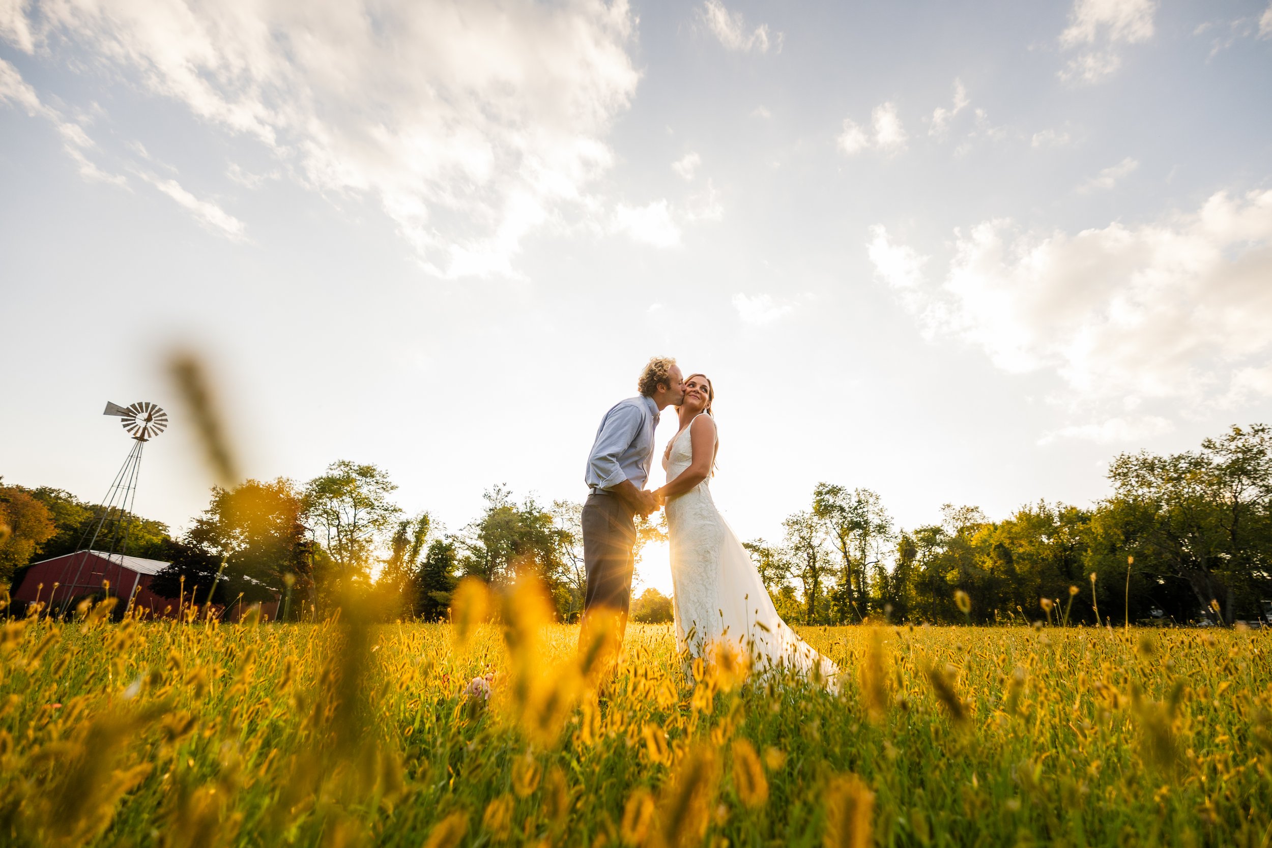 A newlywed couple standing close in a field of yellow flowers during sunset, with trees and a windmill in the background.