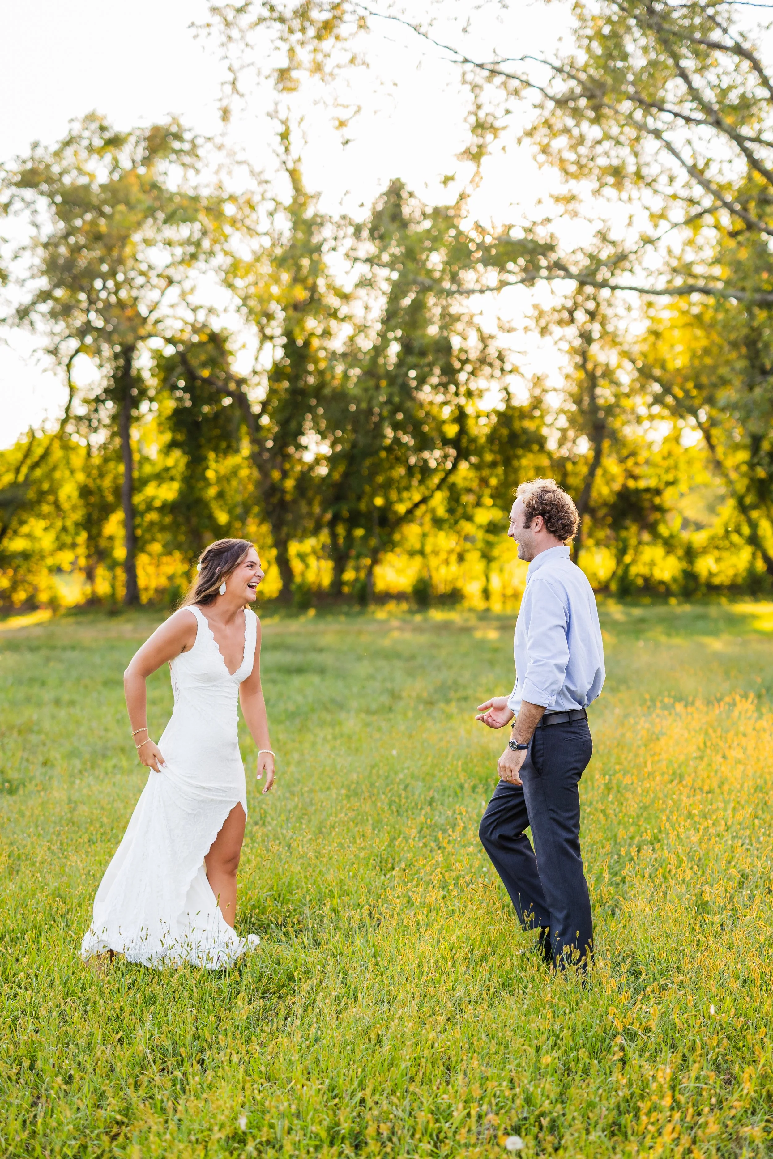 A woman in a white wedding dress and a man in a light blue shirt with dark pants are smiling and laughing in a grassy field with trees in the background during sunset.