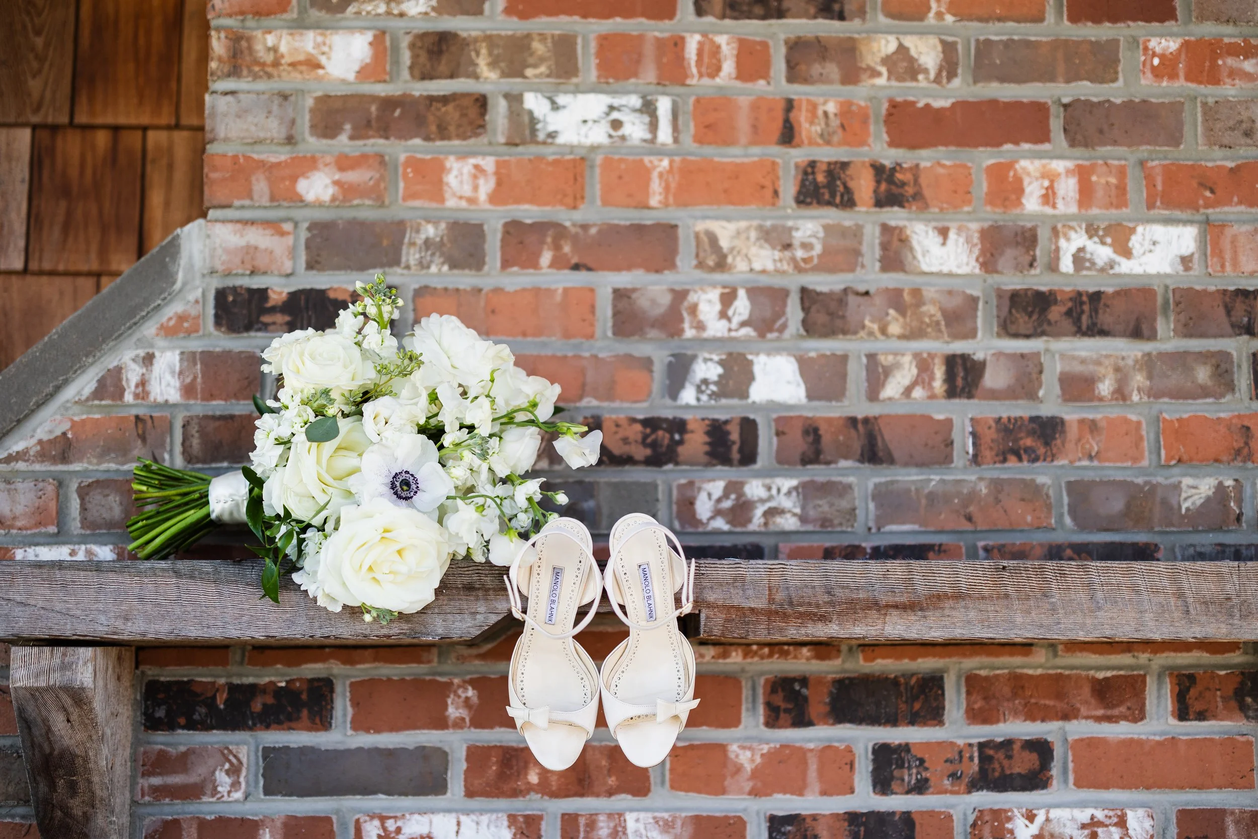 A bridal bouquet of white roses, lisianthus, and an anemone flower resting on a wooden bench, with a pair of white high-heeled sandals hanging beside it, against a brick wall background.