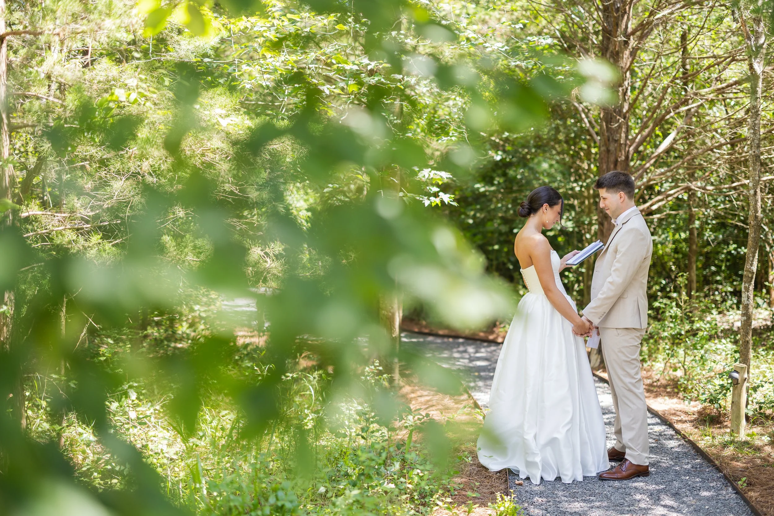 A bride and groom holding hands while exchanging vows in a forested area during a wedding ceremony, with a lush green background and sunlight filtering through the trees.