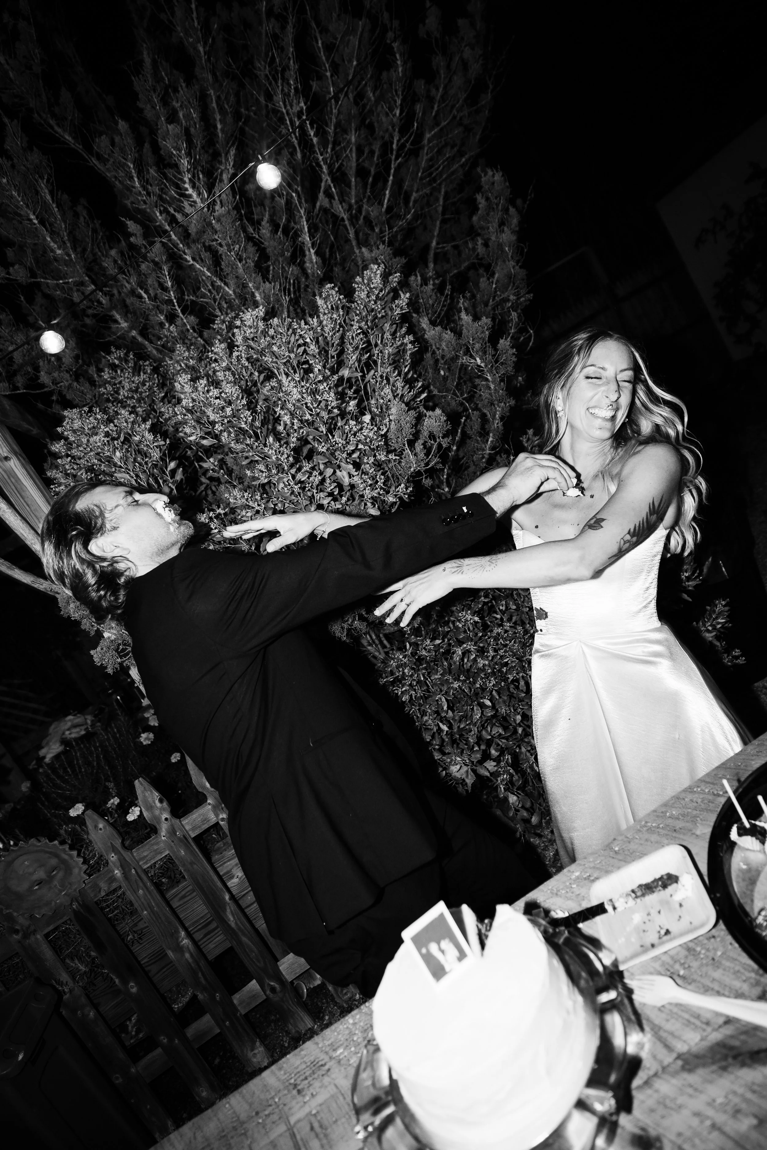 A couple at a wedding reception, the groom in a black tuxedo playfully pulling at the bride's dress, both smiling and laughing, in an outdoor setting with string lights and greenery.